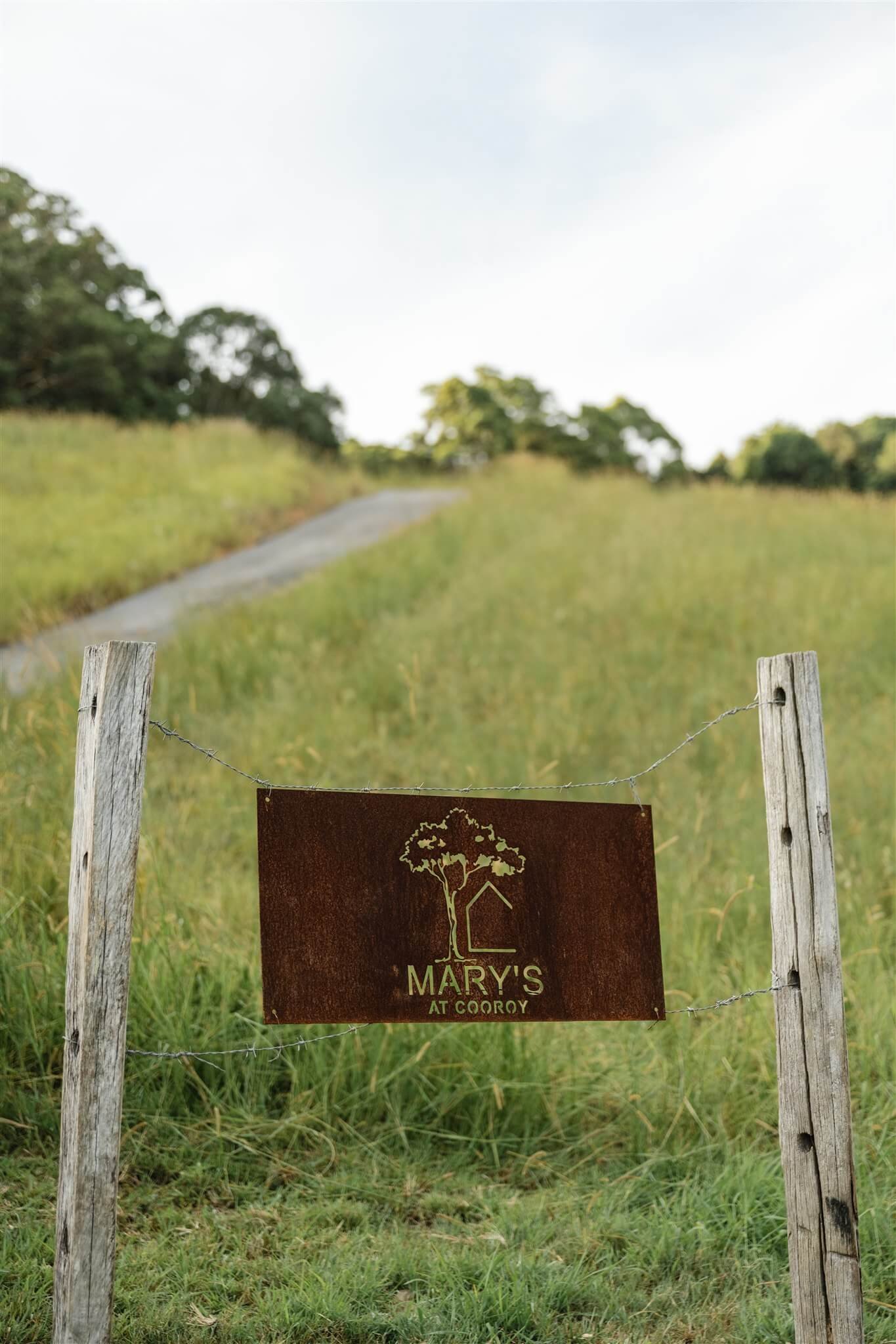 A rustic wooden fence with a sign that reads 'Mary's at Cooroy' hangs on wire across the posts, with a grassy hill and trees in the background.