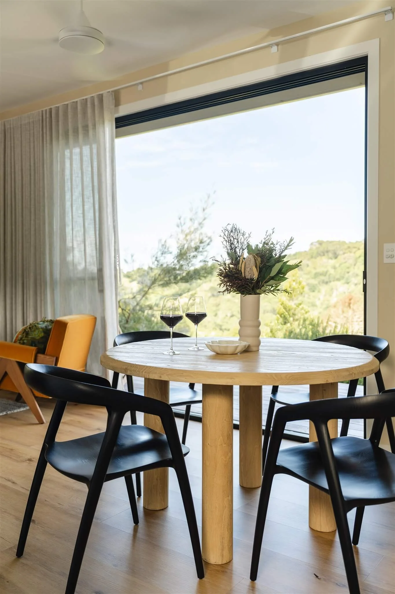 A dining area with a round wooden table, four black chairs, a vase with flowers, two glasses of red wine, and a large window showing a view of trees and hills.