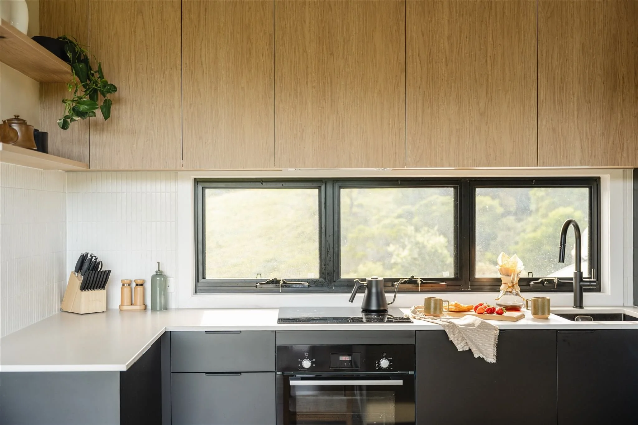 Modern kitchen with black lower cabinets, white countertop, and light wooden upper cabinets. A window with a mountain view behind the countertop. On the counter, there is a black kettle, a tray with fruit, two cups, and a paper drip coffee maker. To 