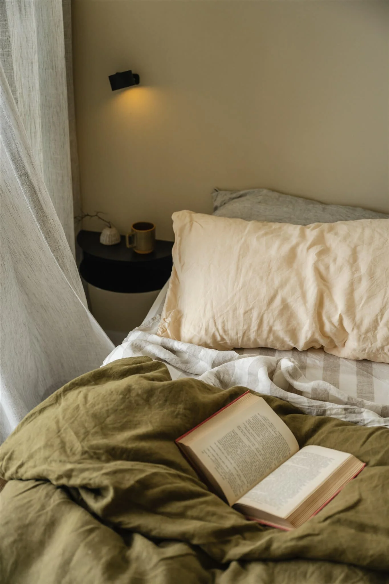 A cozy bedroom scene shows a bed with beige, gray, and white bedding, a nightstand with a cup and decor, and a wall-mounted reading light in warm lighting. An open book rests on a green blanket in the foreground.