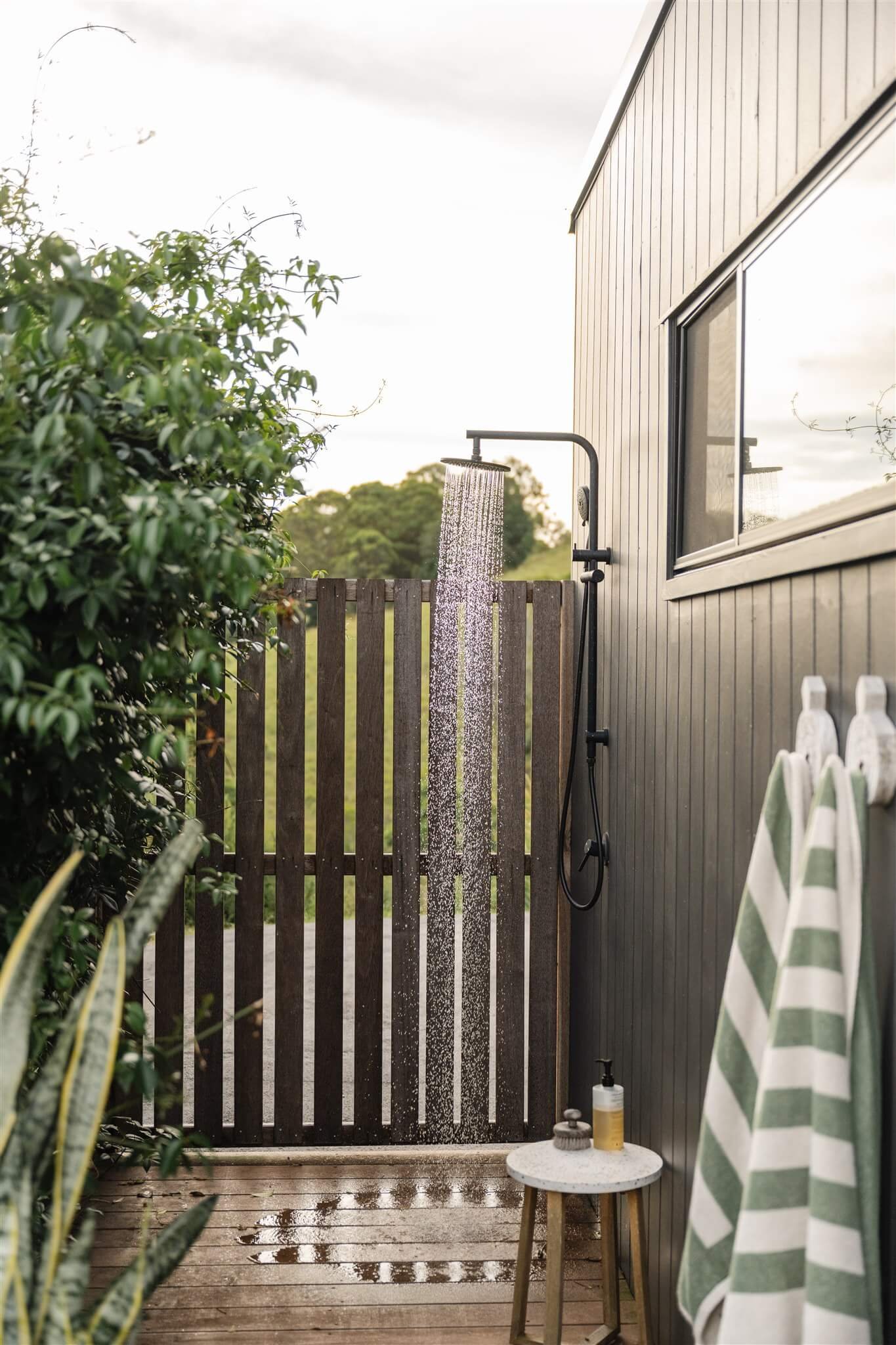 Outdoor shower with water running, positioned against a dark wood fence, with green foliage on the left side, a small round table with toiletries, and striped towels hanging on hooks outside a gray building.