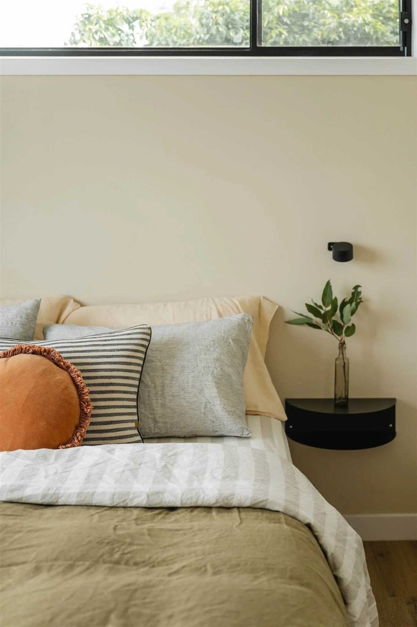 A cozy bedroom corner with a bed featuring multiple pillows, including a round, rust-colored pillow with a ruffled edge. A black floating nightstand holds a clear glass vase with green leafy branches, and a black wall-mounted light fixture is above. 