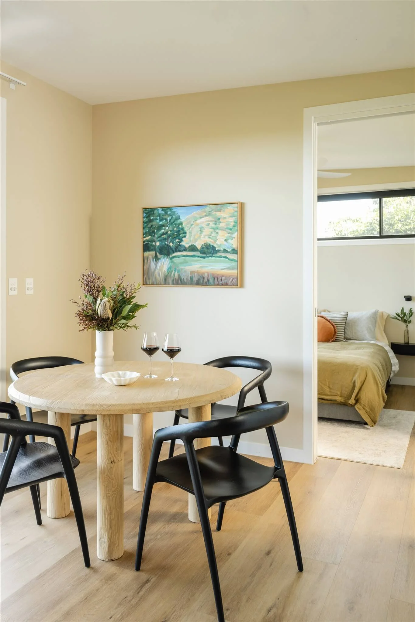 A dining room with a round wooden table, four black chairs, a vase with flowers, and two glasses of red wine, with a bedroom visible through an open doorway.