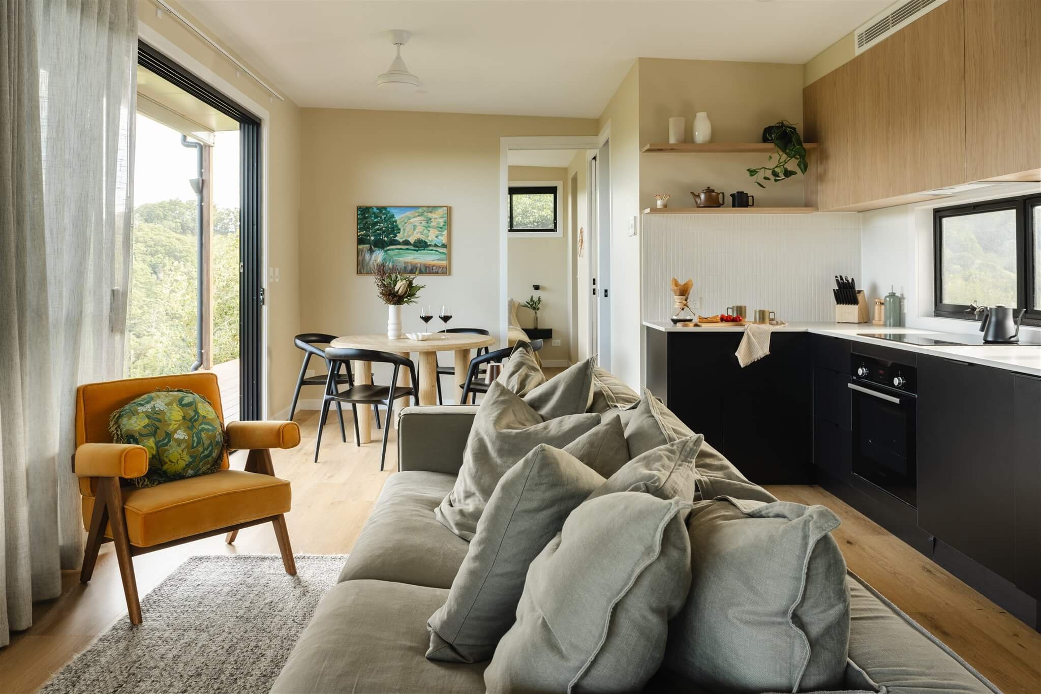 Living room and dining area with beige sofa, a mustard armchair, a round dining table with black chairs, and a view of a lush green outdoor area through sliding glass doors.