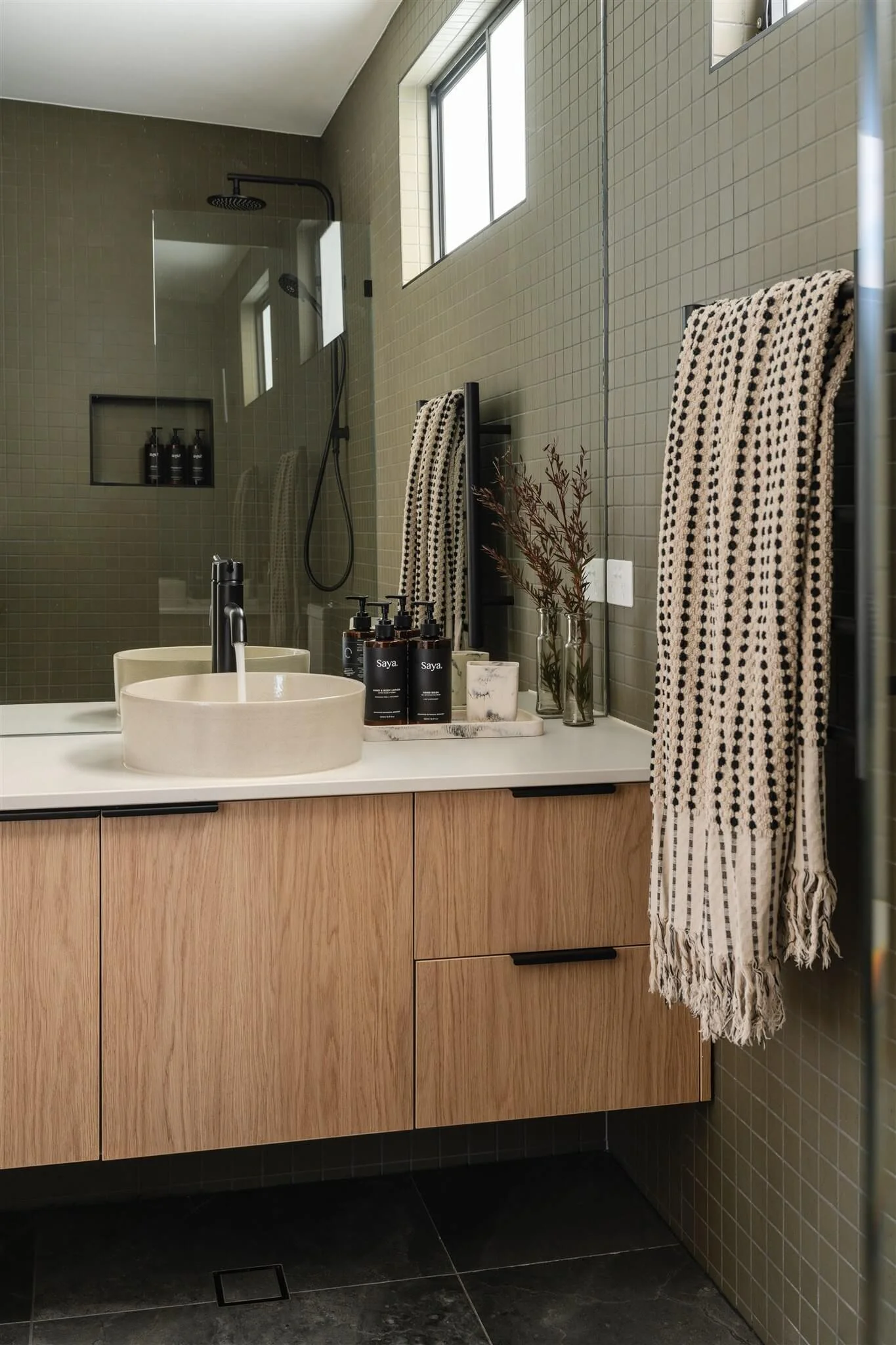 Modern bathroom with wooden vanity, beige round vessel sink, black faucet, wall-mounted black towel rack with a fringed towel, black soap dispensers, and a shower with a glass wall and black shower fixtures. There are small windows on the tiled wall.