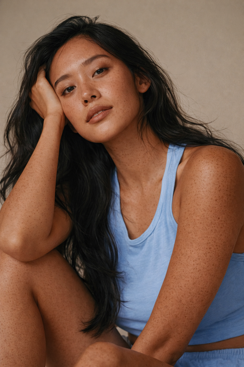 A woman with long black hair, light brown skin with freckles, wearing a light blue tank top, resting her head on her hand and looking at the camera.