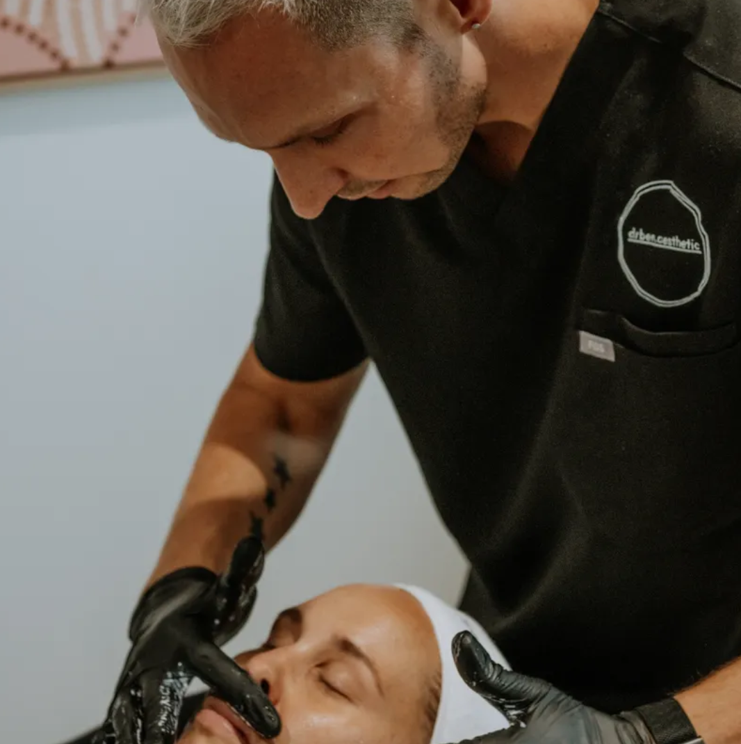A person with a tattoo on their arm is giving a facial to another person who is lying down with their eyes closed, wearing a headband, inside a spa or salon.