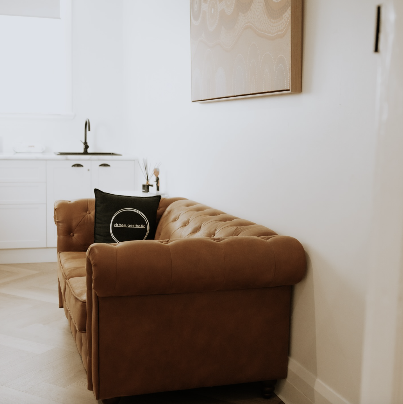 A living room with a brown leather tufted sofa, a black pillow with white text, a small white cabinet, and a painting on the wall.