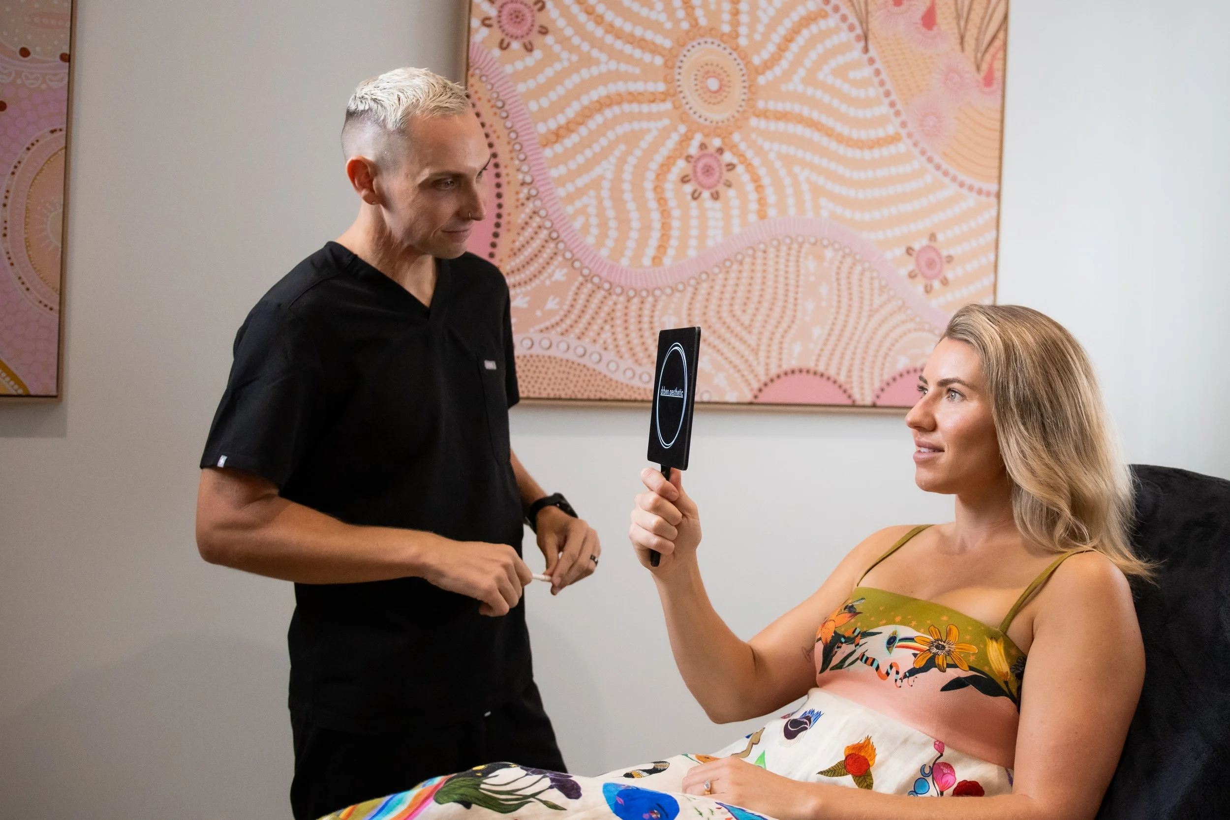 A woman lying in bed with a colorful floral dress holding a mirror, looking at a man standing beside her in a black medical uniform, in a room with pink patterned artwork on the wall.