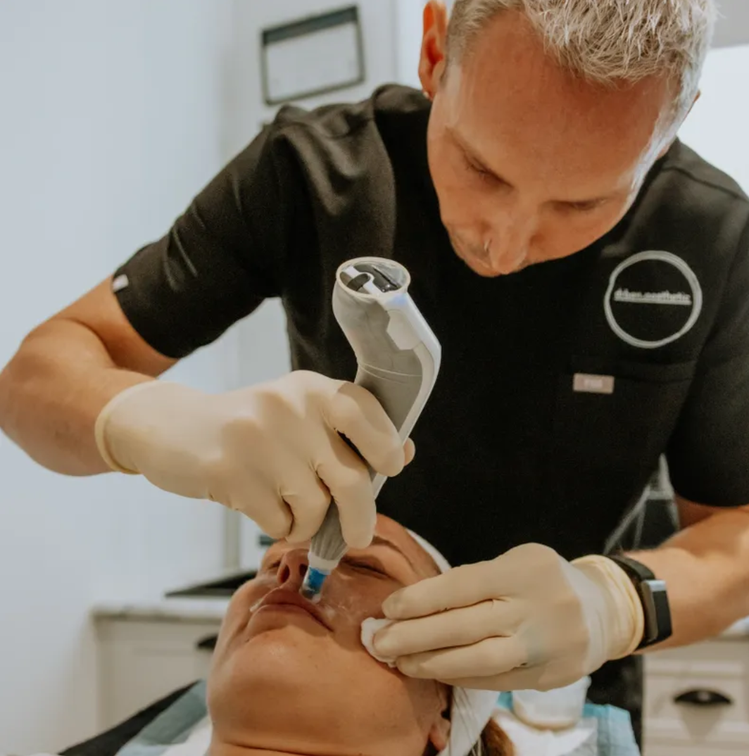 A medical professional wearing gloves and a black uniform administering a cosmetic treatment to a woman's face using a handheld device.