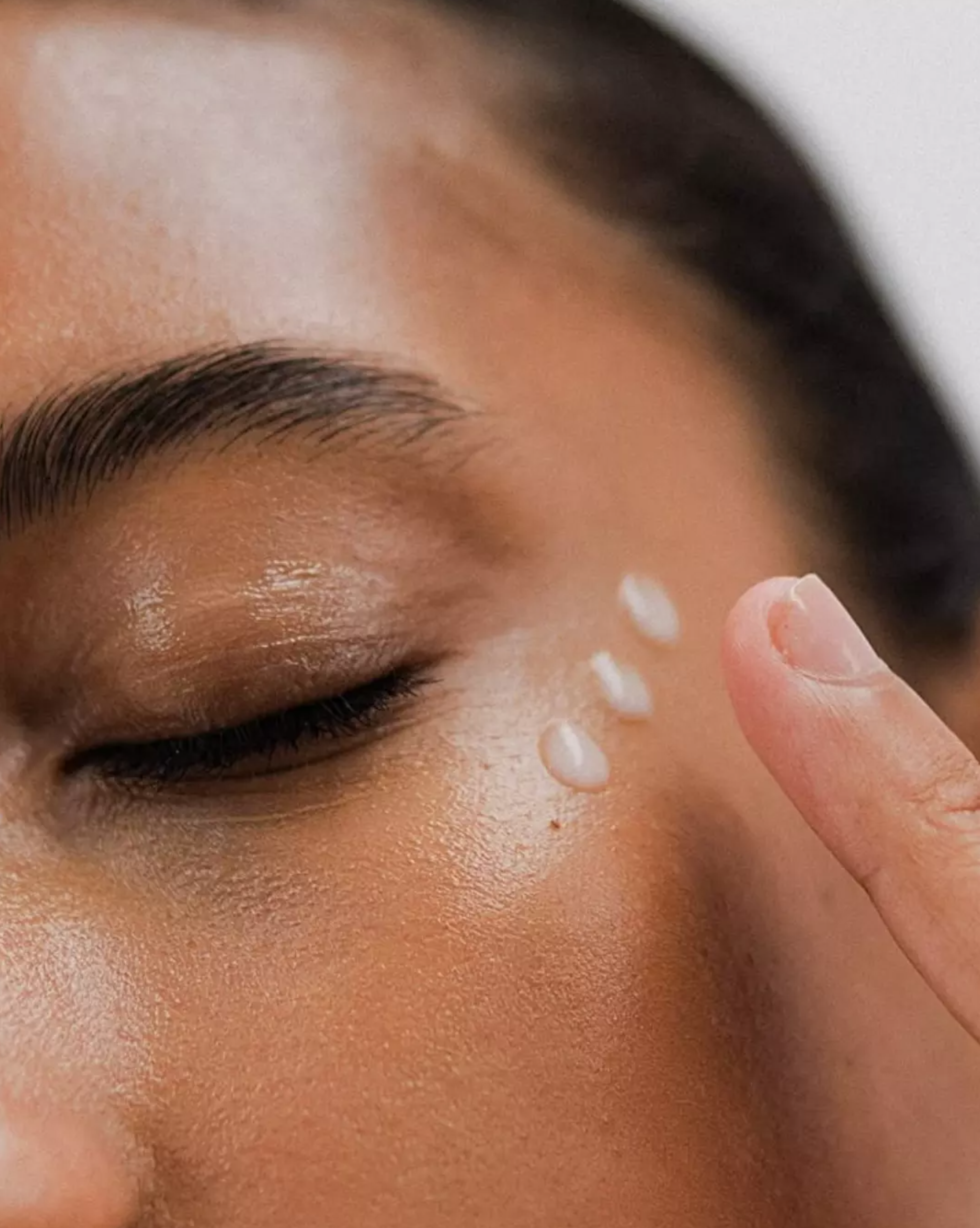 A close-up of a person's face with three drops of a white liquid on their cheek, with their finger next to the drops.