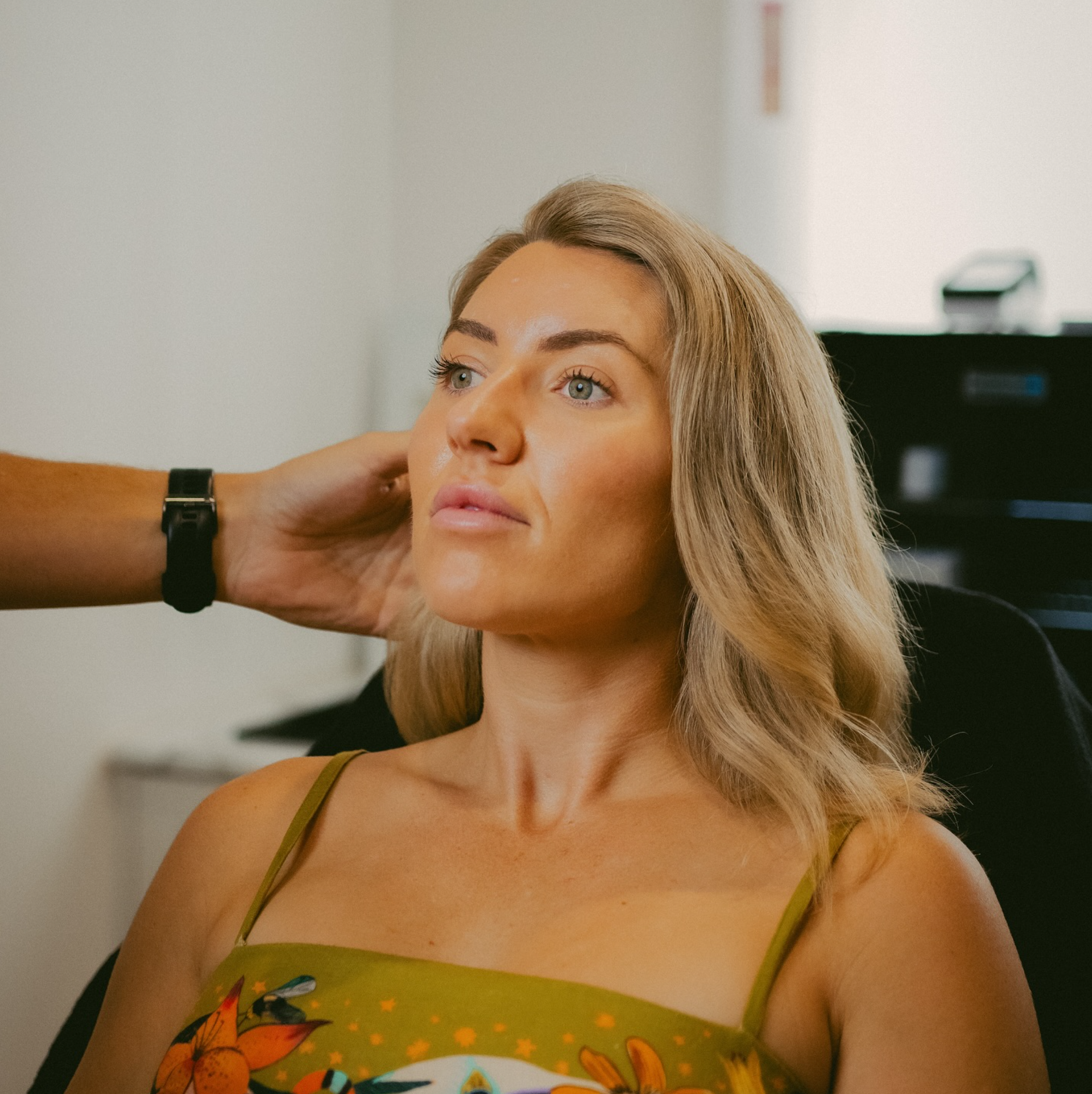 A woman with blonde hair and blue eyes looking slightly upward while sitting in a chair. A person's arm is gently touching the back of her head.
