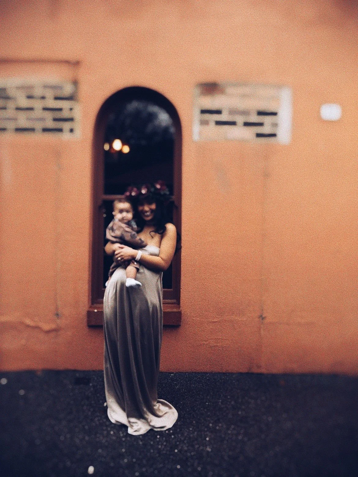 A woman dressed in a long gown holding a young girl, standing in front of an orange-colored wall with two vents and a window with a person visible through it at night.