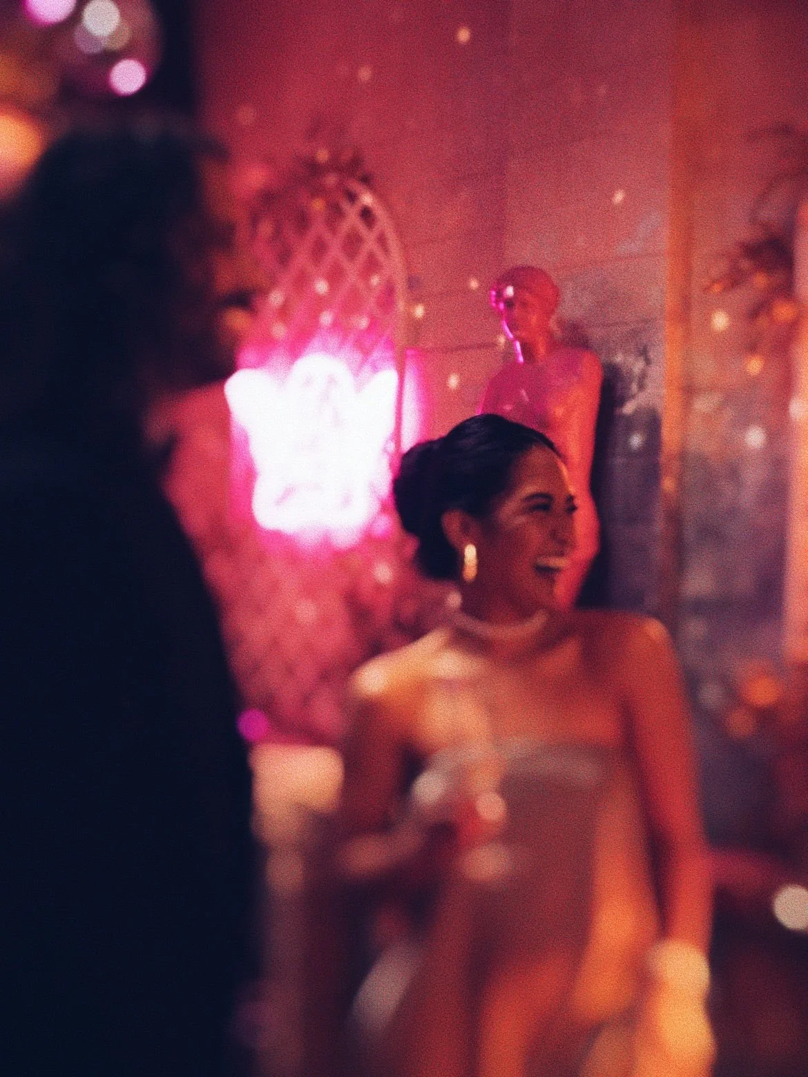 A woman in a strapless dress laughing and talking to a person whose face is not visible, in a dimly lit, colorful party or nightclub setting with neon lights and a statue in the background.