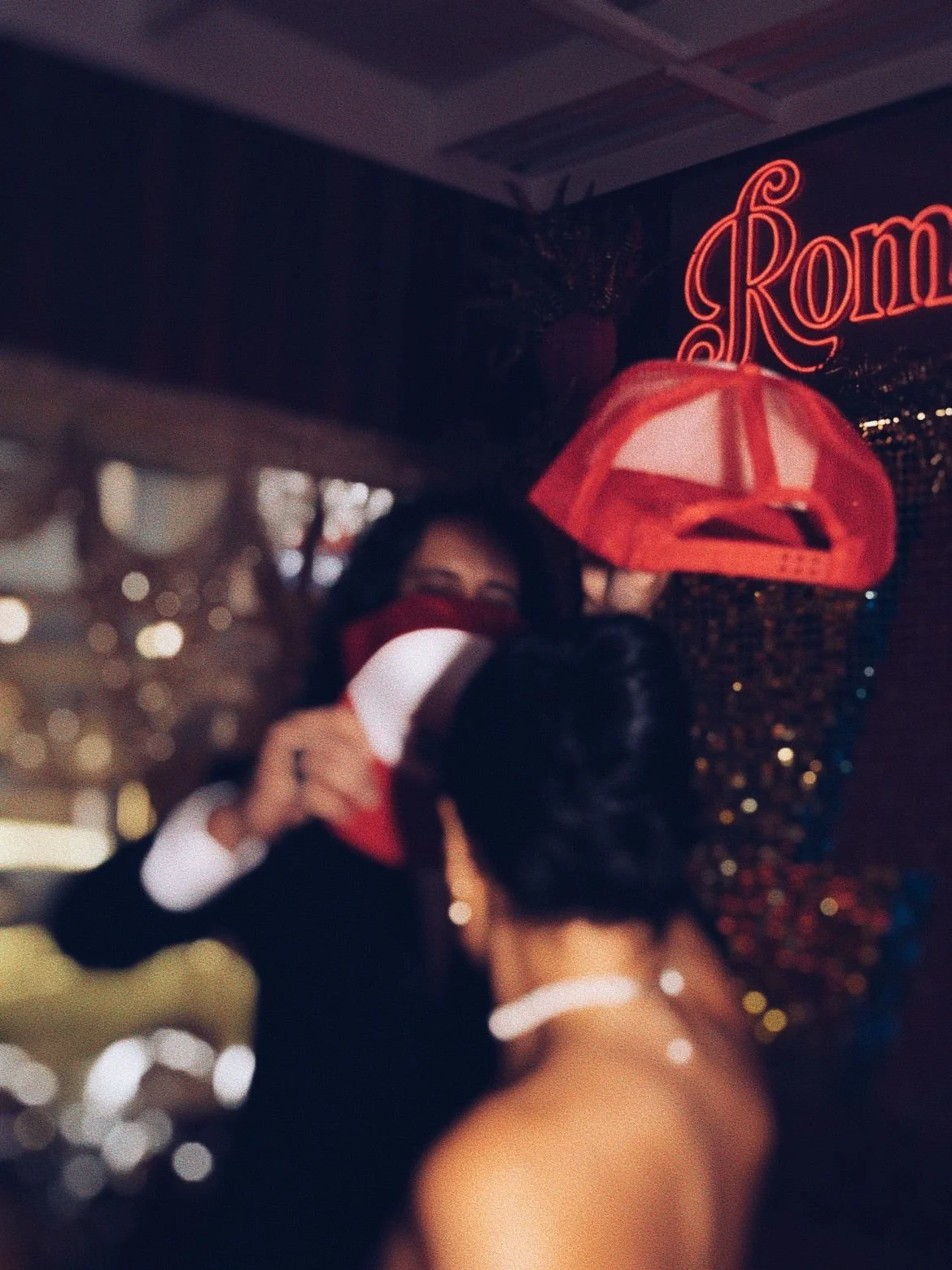 Two women at a festive event, one adjusting a Santa hat on the other's head, with a neon sign saying 'Roma' in the background.