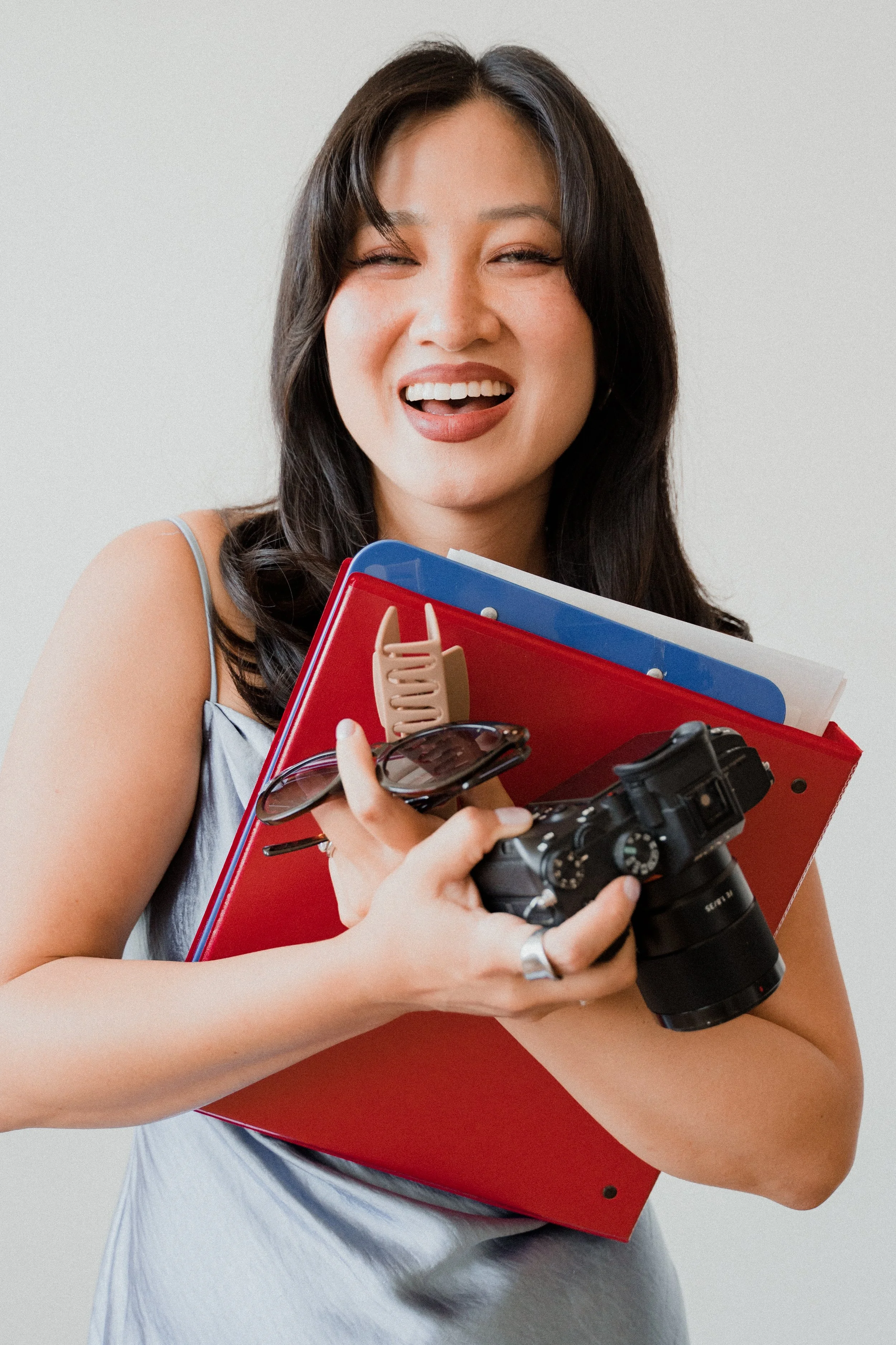 A woman smiling and holding a camera, a red folder, and other items, with a light gray background.