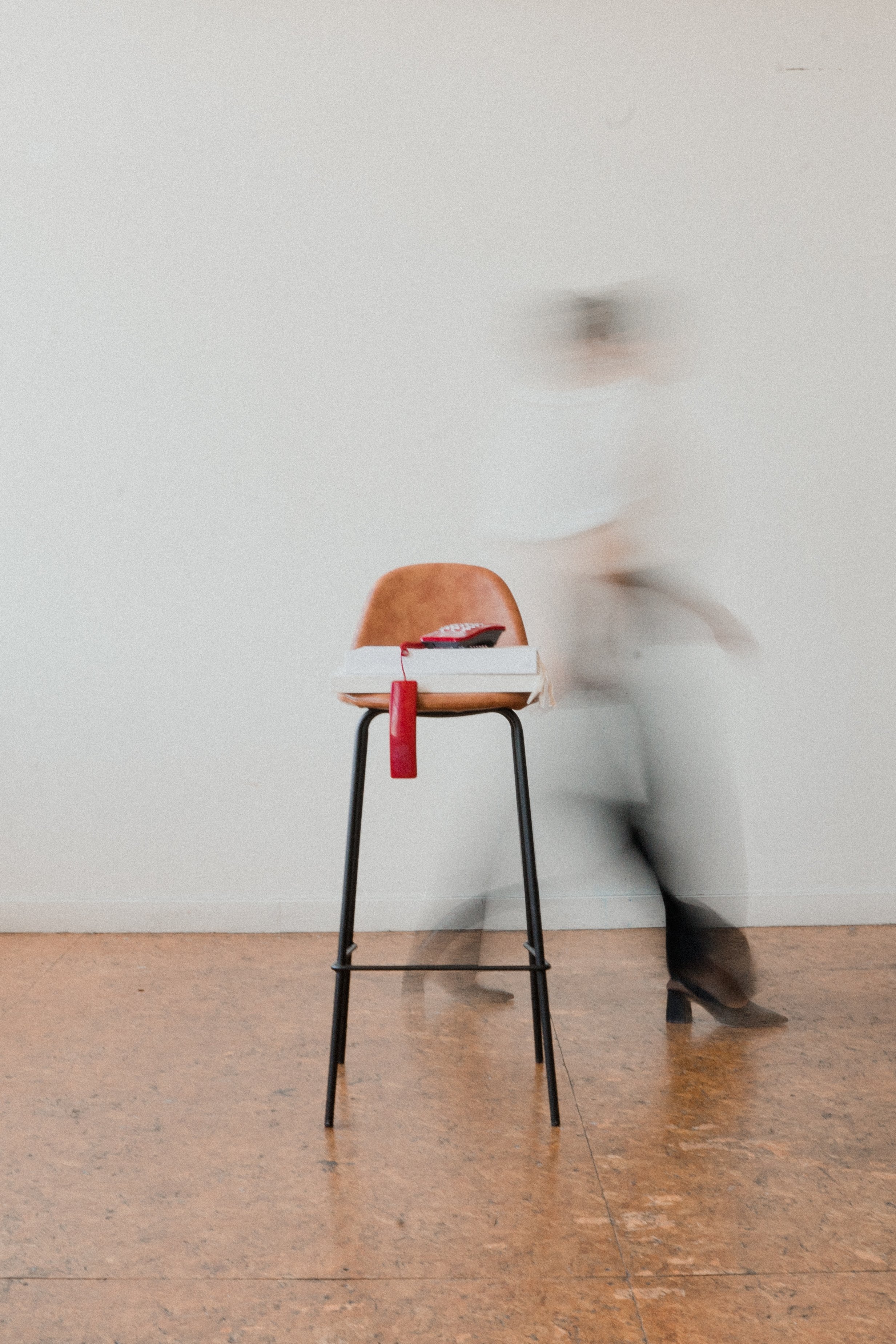 A wooden chair with black metal legs in the center of a room. On the chair sits a stack of papers, a red bookmark, and a black object that looks like a small pouch. To the right of the chair, a person is blurred in motion, walking past the chair. The background features a plain, light-colored wall and a hardwood floor.