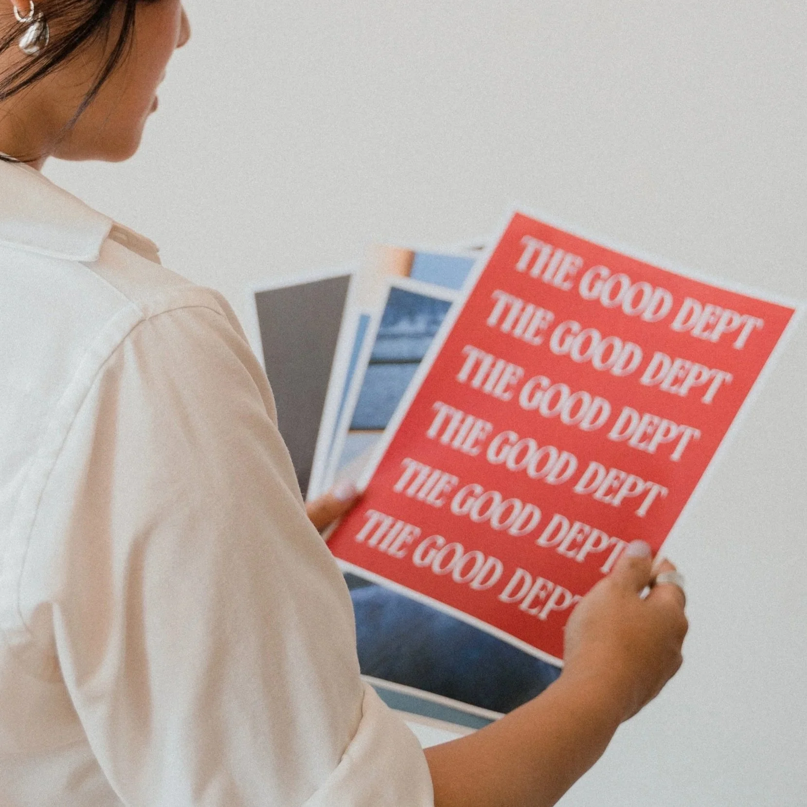 A person in a beige shirt is holding a red flyer with white text that reads 'THE GOOD DEPT' multiple times. The person's face is not visible, and they are standing indoors with a white wall in the background.