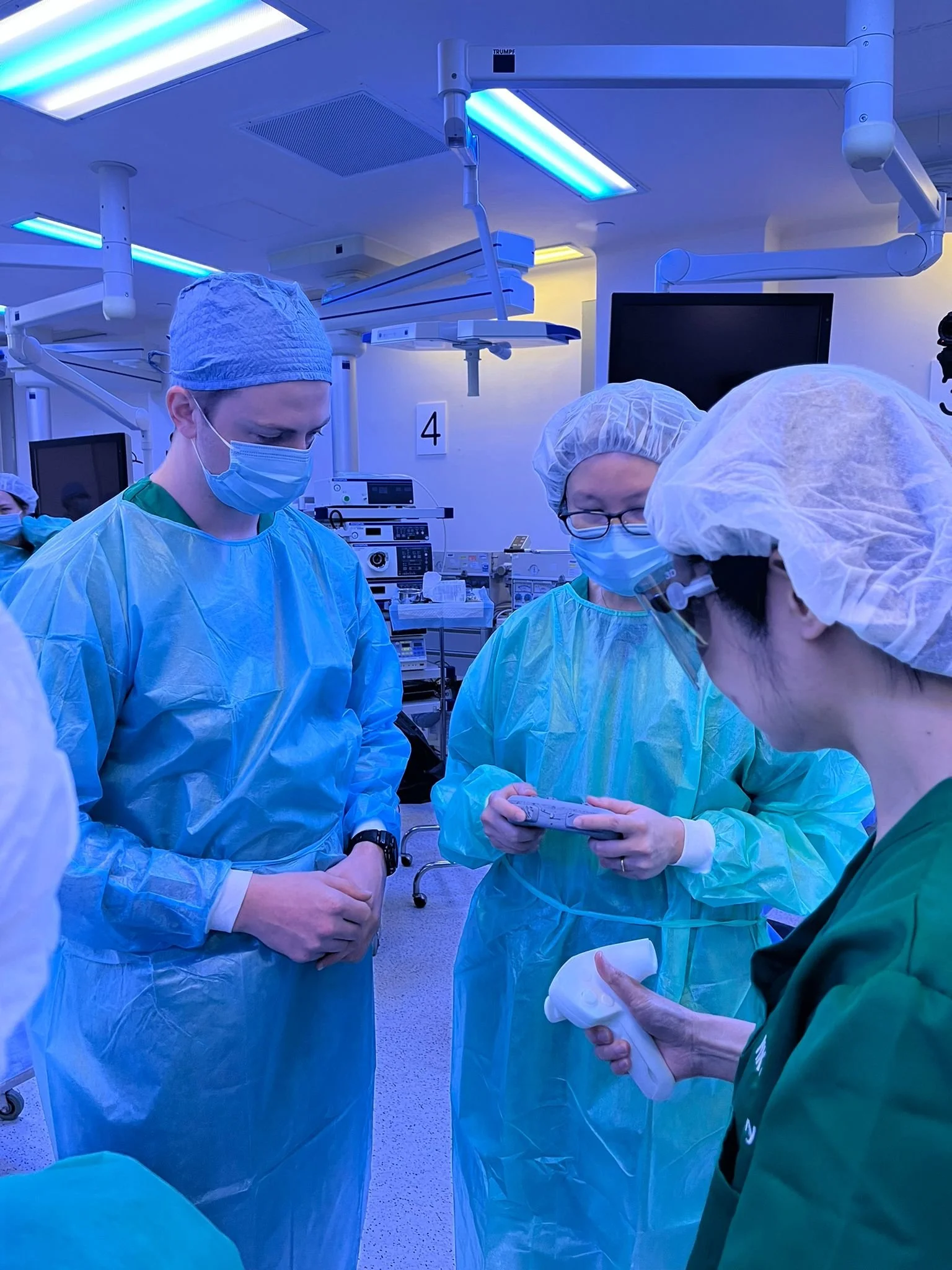 Medical team in operating room wearing surgical scrubs, masks, and hair covers, gathered around a procedure table, with medical equipment and monitors in the background.