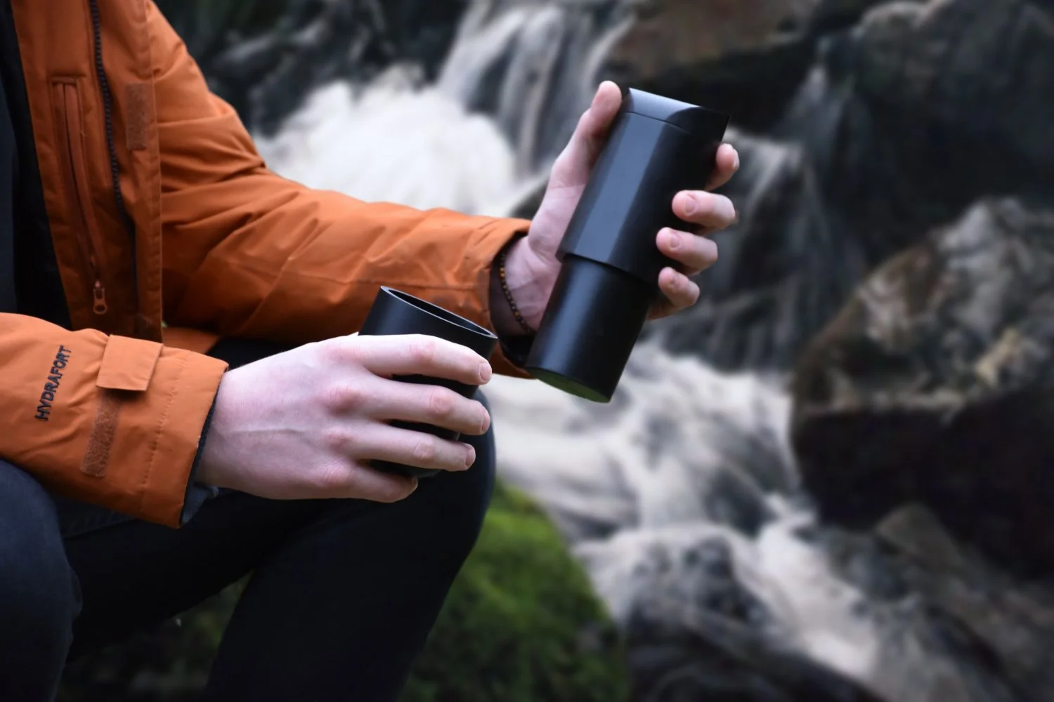 A person in an orange jacket holding a black thermos and a cup in front of a waterfall.