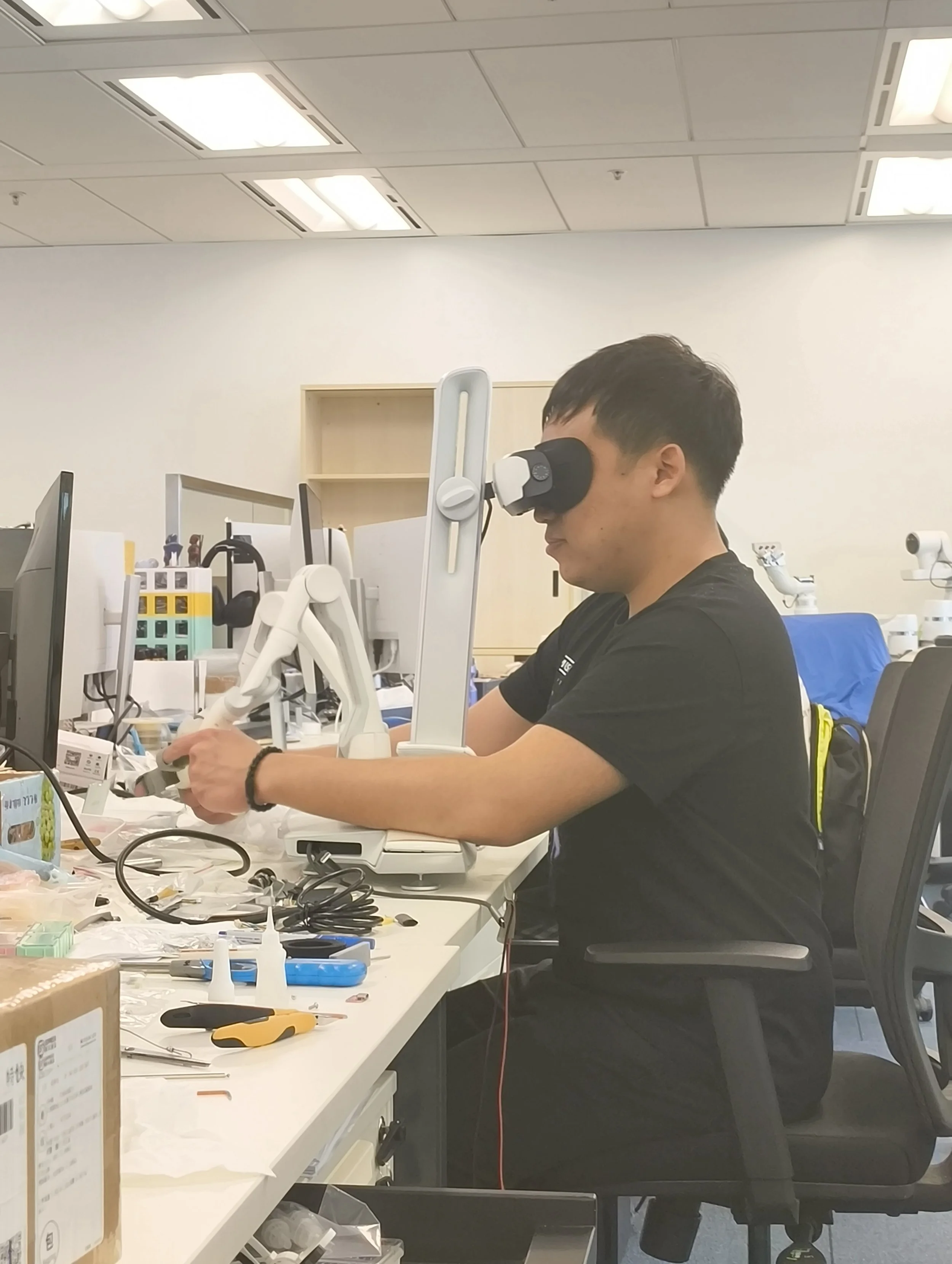 A man wearing a black shirt and a wristwatch is sitting at a table in an office or laboratory, using a microscope. The table is cluttered with various tools, electronic devices, and supplies. In the background, there are desks, computer monitors, shelves, and office lighting.