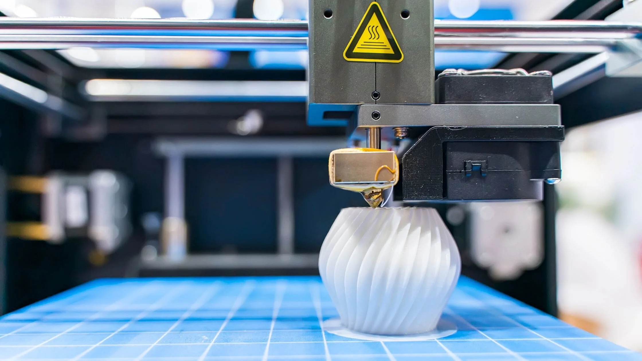 A 3D printer printing a white, ridged, spherical object on a blue grid surface.