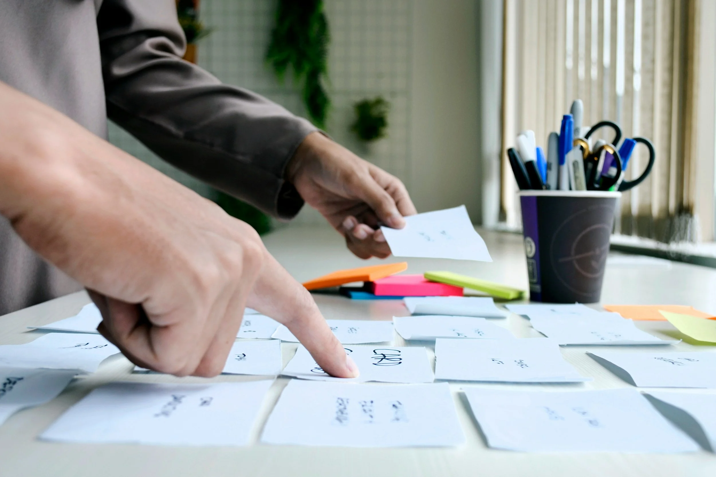 Close-up of hands arranging small notes on a table, with a cup of pens and bright background with curtains.
