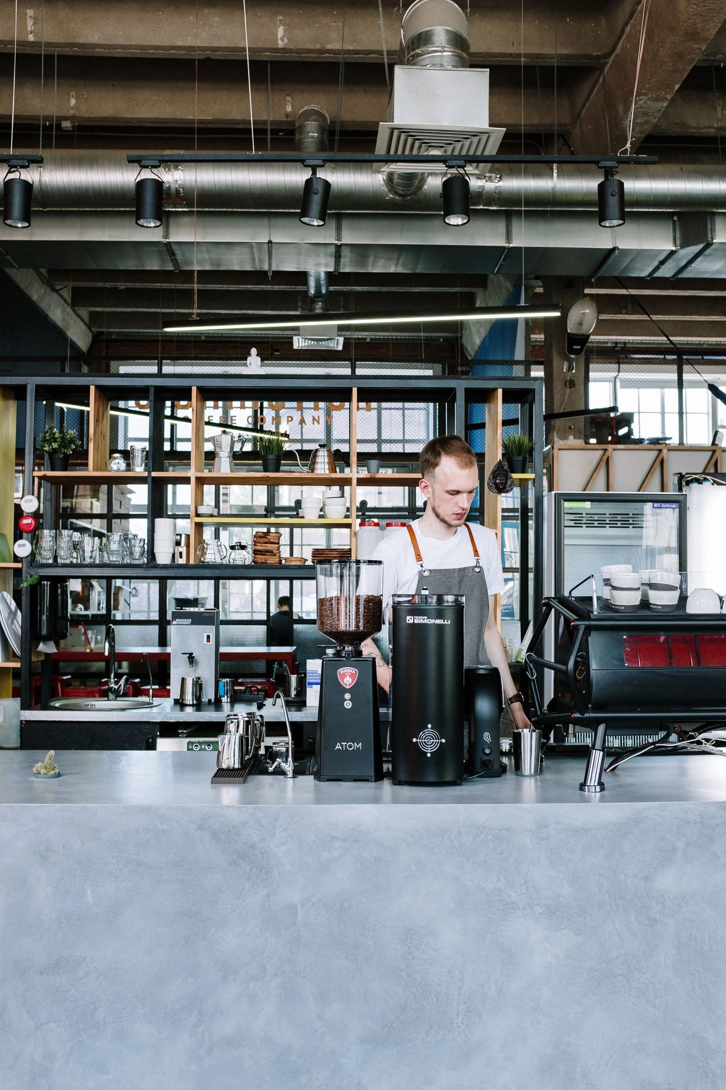 A barista preparing coffee behind a counter in a modern, industrial-style coffee shop with exposed ductwork and large windows.