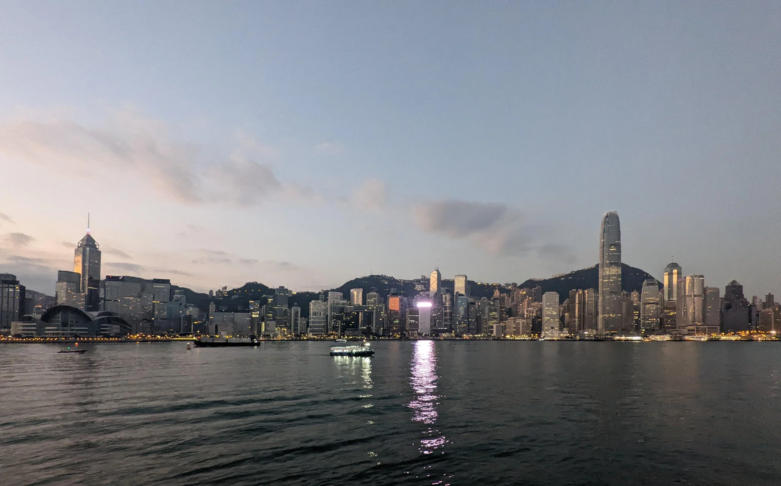 Hong Kong skyline view across the harbour at dawn with high-rise buildings, including a tall, sleek building and a tower with red and white lights, reflecting on the water.