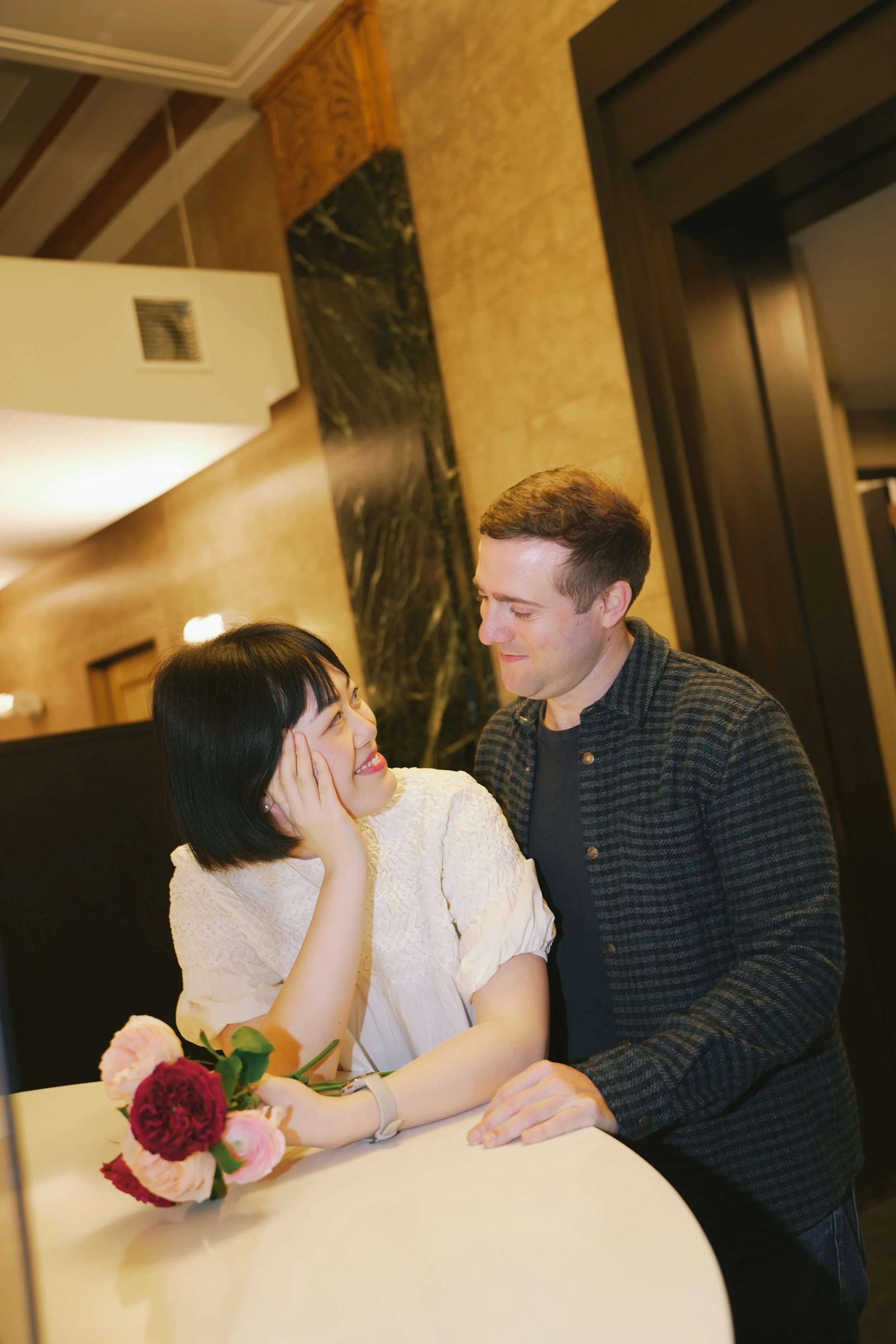 A man and a woman share a moment of affection at a table, with a bouquet of flowers in front of her, in an elegant indoor setting.