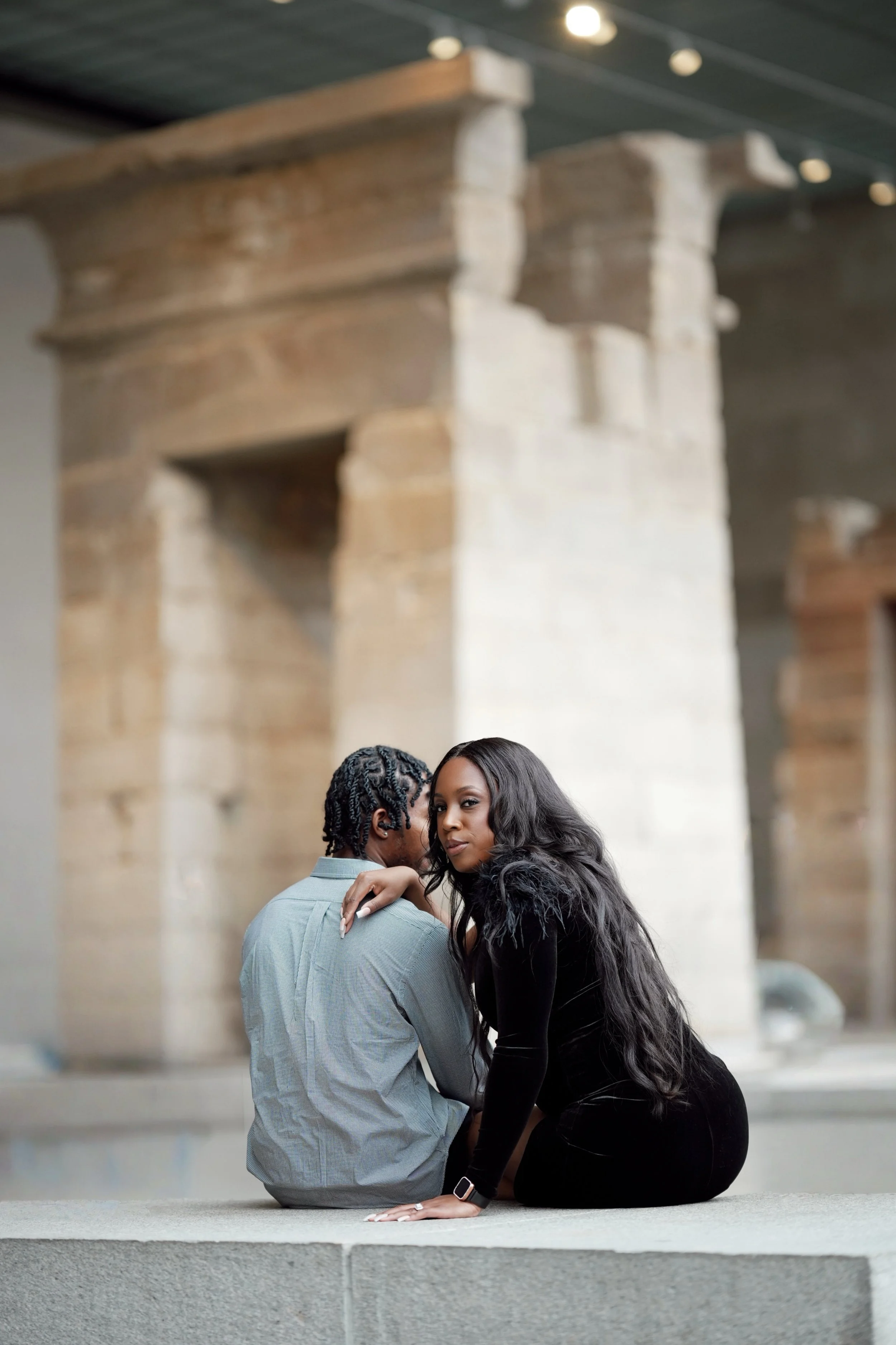 A woman with long black hair and a black dress sitting on a concrete surface, leaning close to a man with dreadlocks and a plaid shirt, in front of ancient ruins or stone structure.