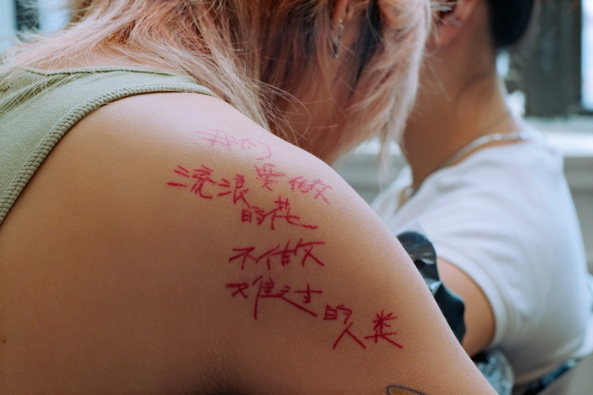 Close-up of a person's shoulder with red handwritten Japanese characters on the skin, and part of another person wearing a white shirt in the background.