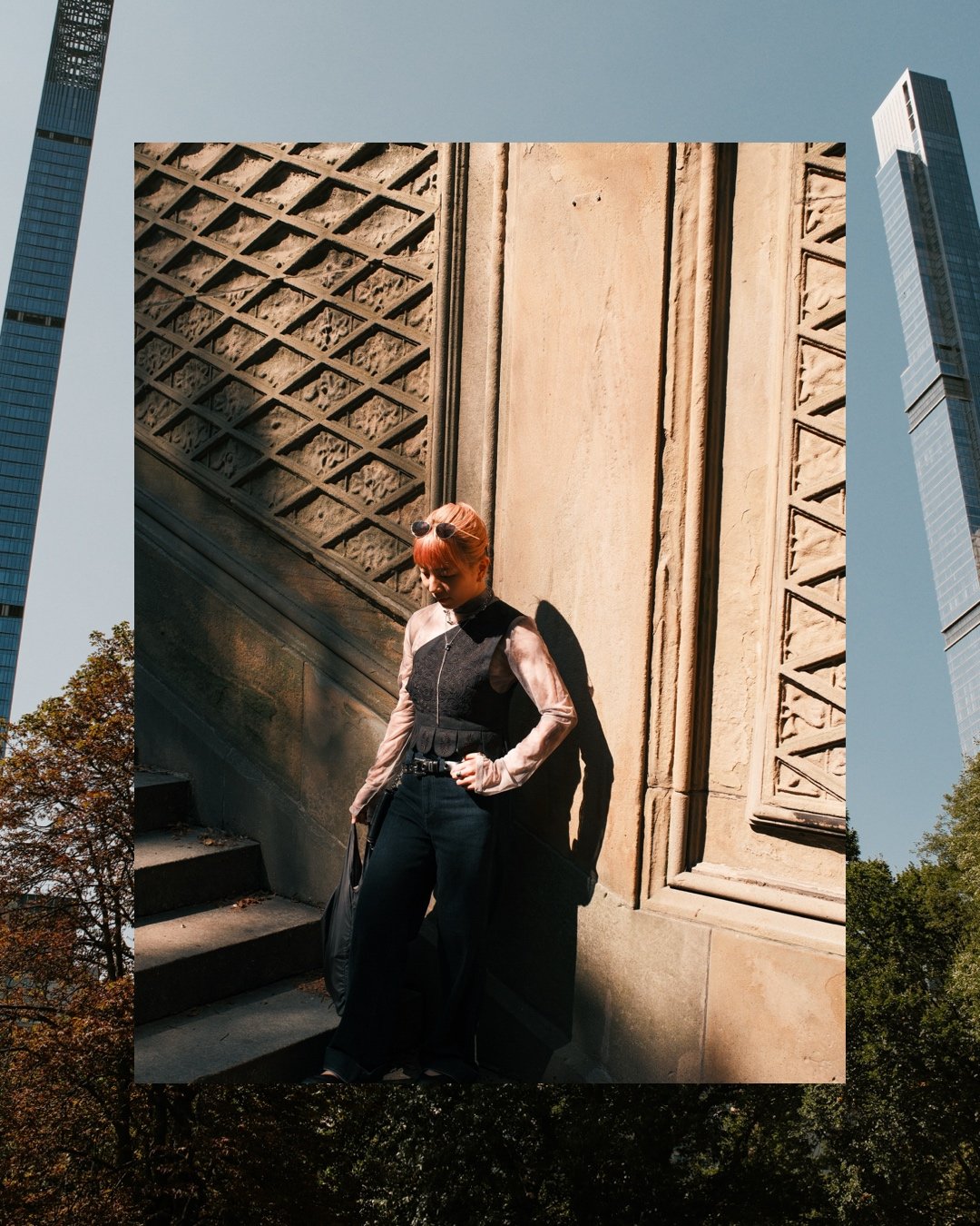 A woman with red hair and sunglasses on her head stands against a stone building, casting a shadow on the wall behind her. She is wearing a black dress with sheer sleeves and black pants, with her hands resting on her hips.