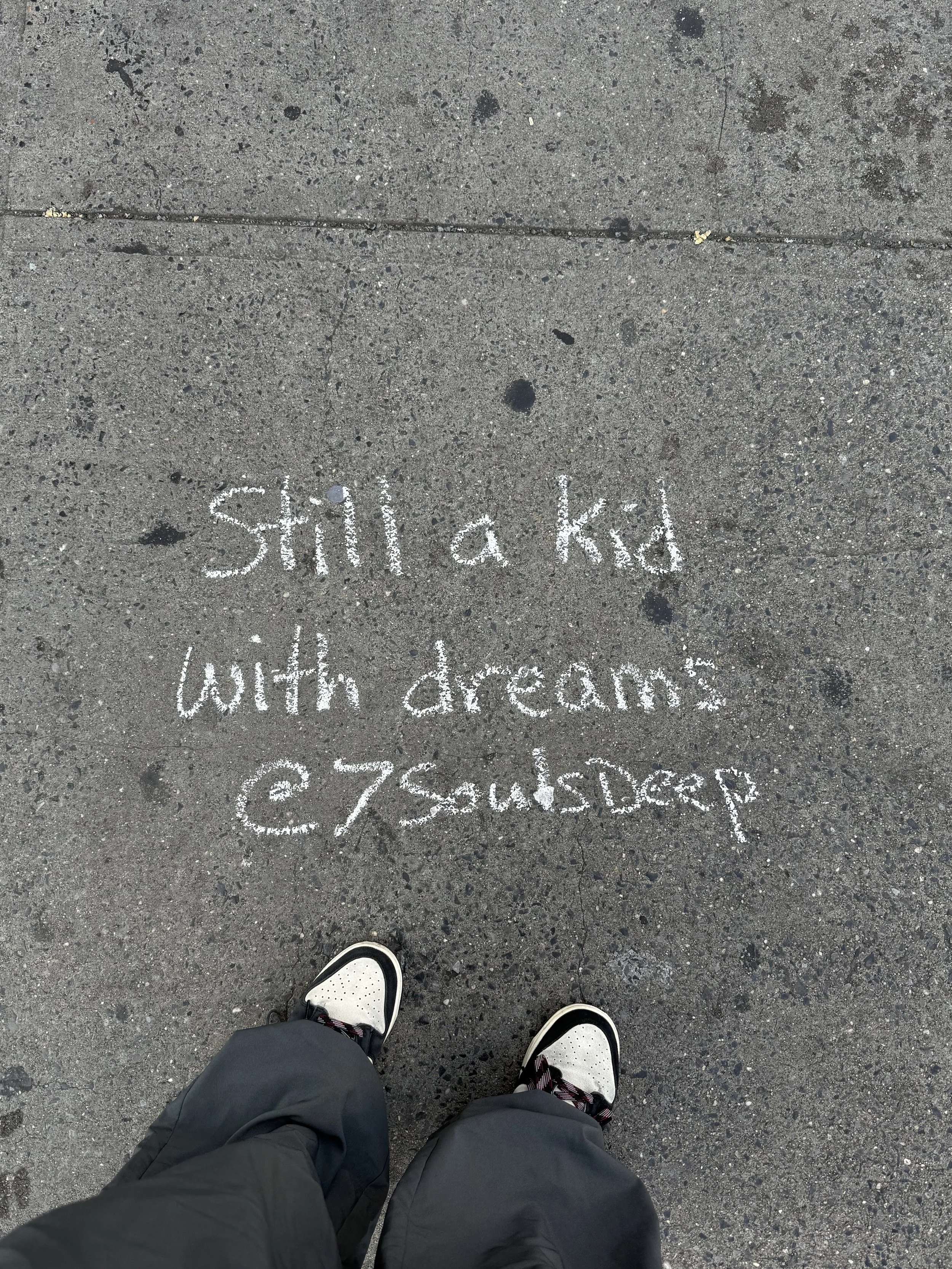 Concrete sidewalk with white chalk writing that says 'Still a kid with dreams' and a pair of black and white sneakers at the bottom of the image.