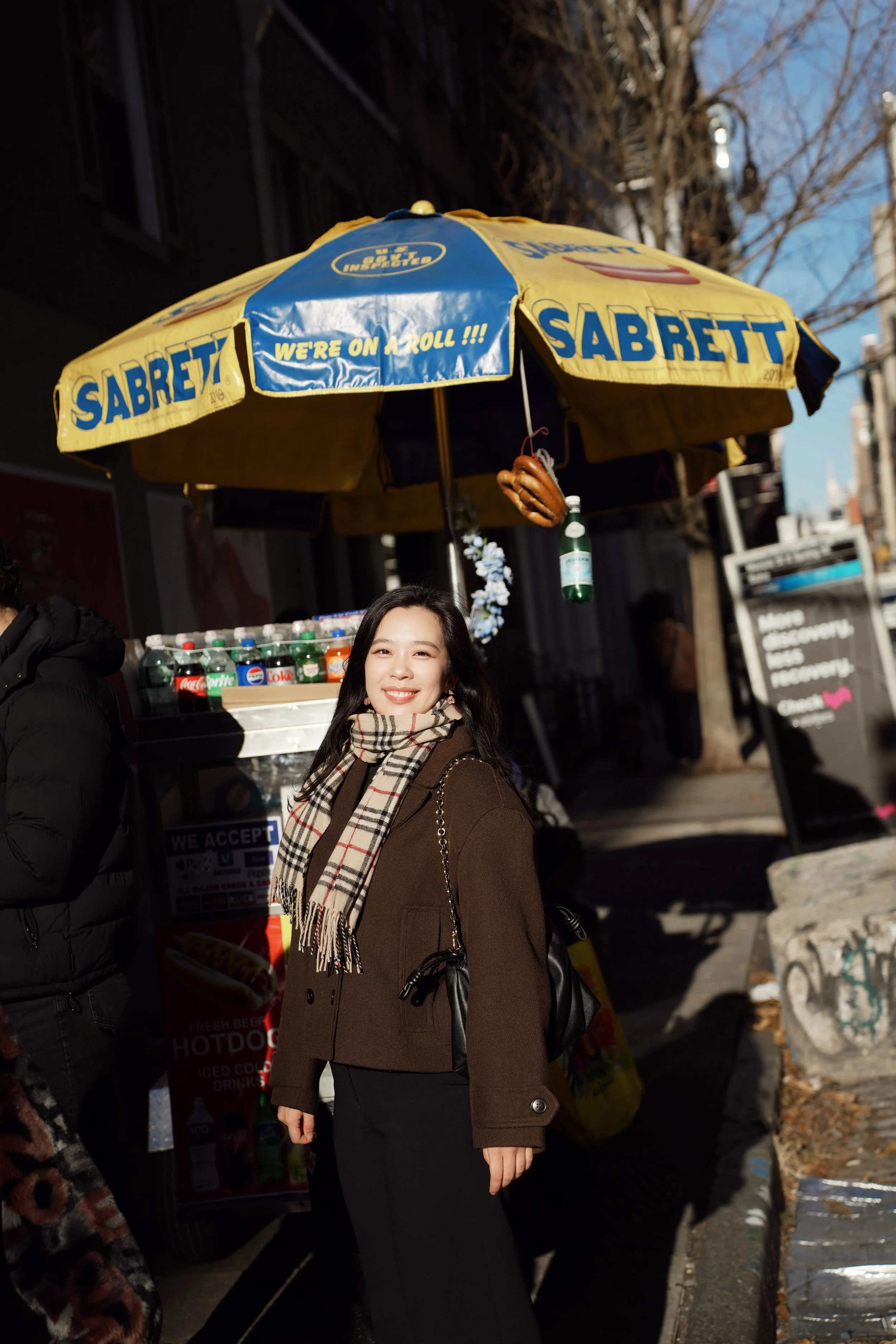A young woman standing on a city sidewalk smiling at the camera under a yellow umbrella with blue writing that says 'Sabrett' and 'We're on A Roll!!!'. She is wearing a brown coat and a plaid scarf, with a black purse over her shoulder. Behind her is