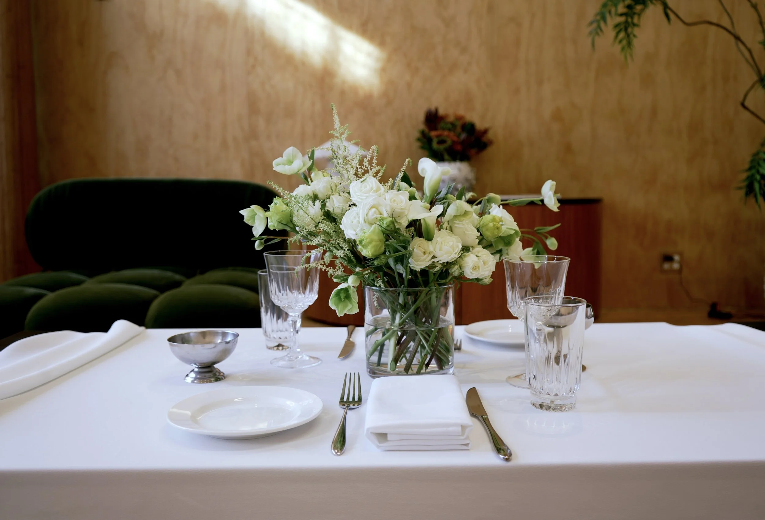 Elegant table setting with a large white floral centerpiece, crystal glasses, a white napkin, and silverware on a white tablecloth, in a room with wooden walls and a dark green velvet sofa in the background.