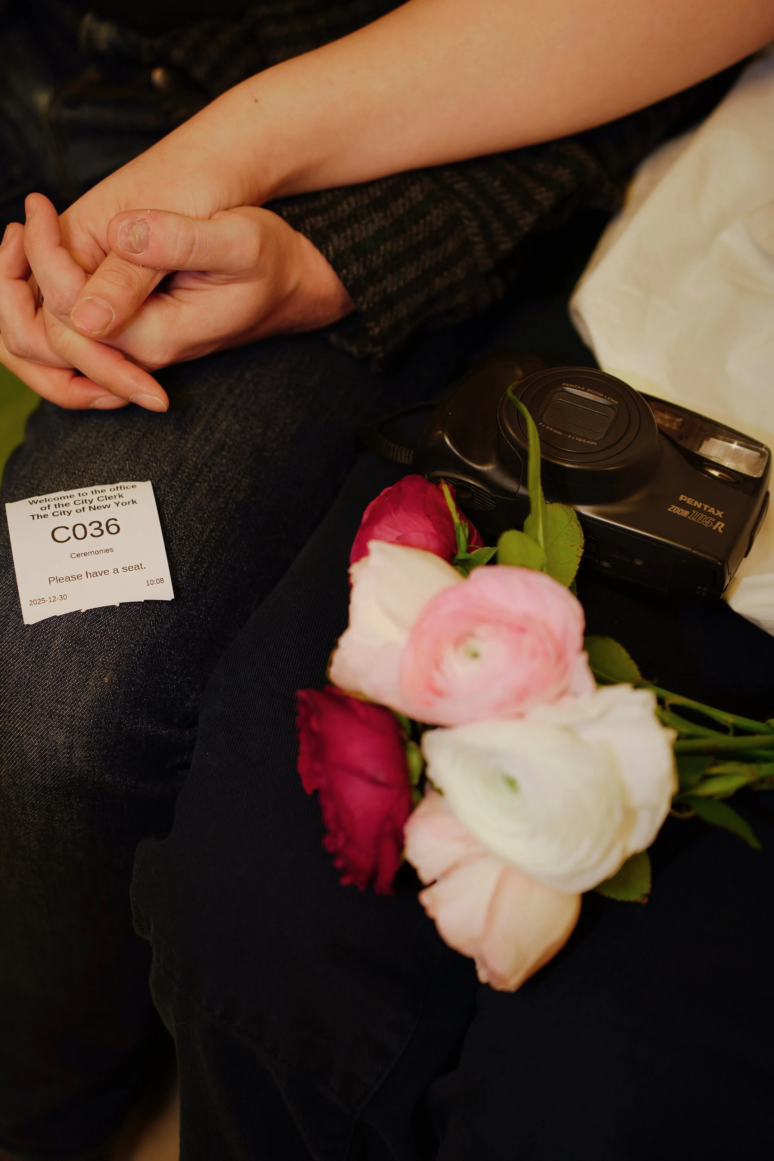 Close-up of two people holding hands, one with a wedding or engagement ring, sitting on a bench with a bouquet of pink and white flowers, a camera, and a ticket stub for an event in New York City.