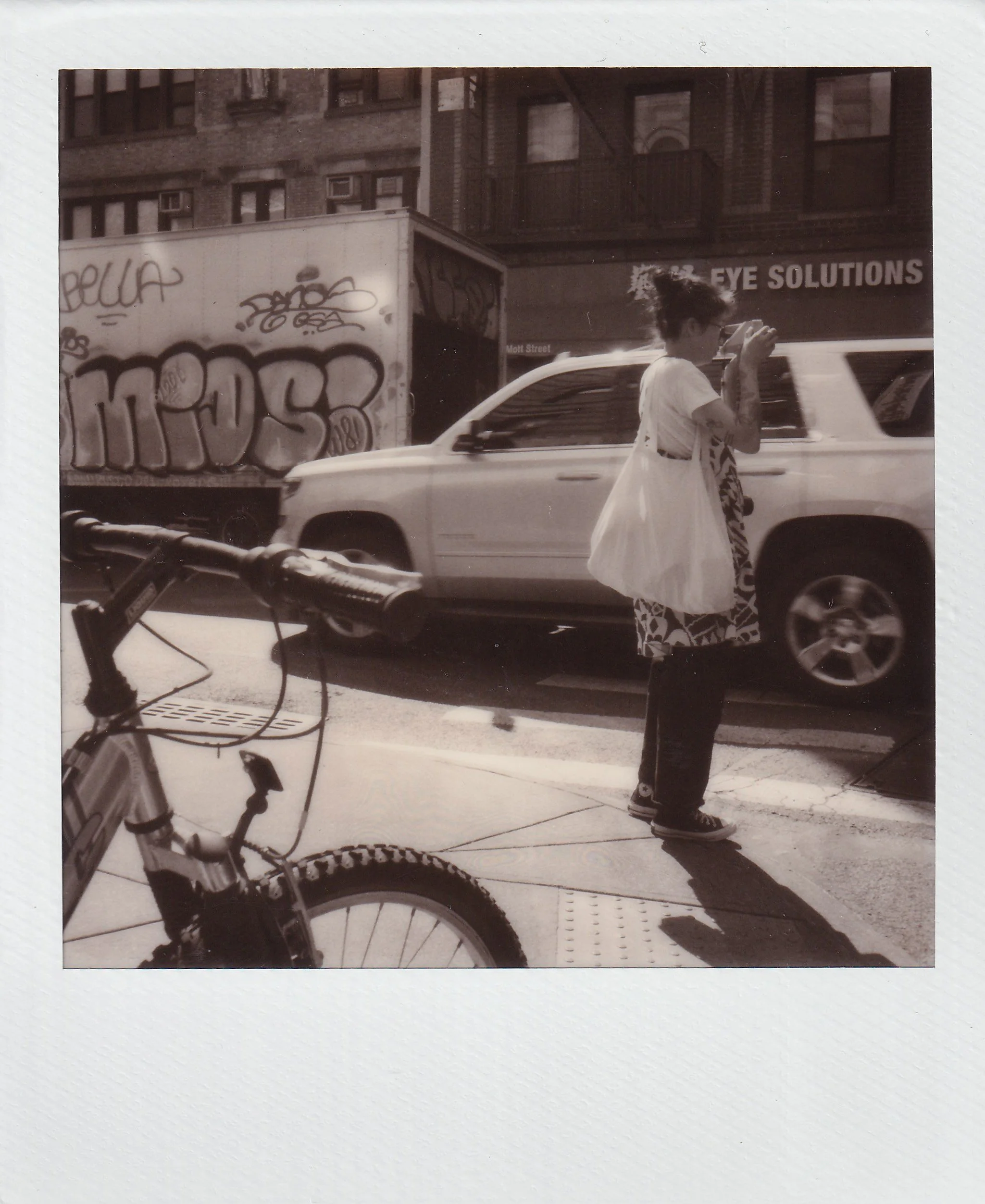 A woman with tattoos on her arms wearing a white top and a patterned skirt, standing on a city sidewalk and looking at her phone. A bicycle is partially visible in the foreground, and a white SUV is parked behind her. Graffiti is on a truck in the background, along with buildings and a sign for an eye solutions shop.