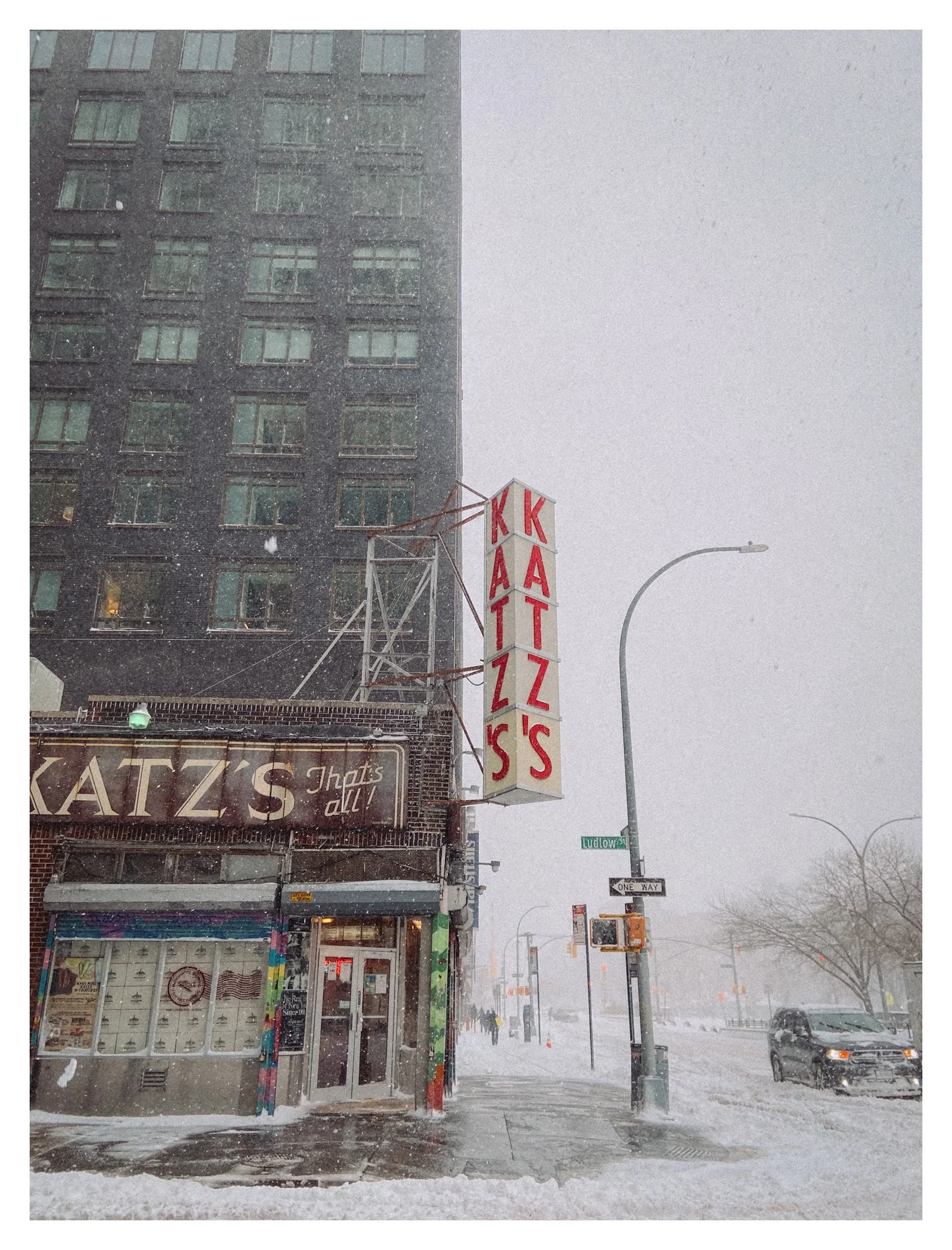 Snow falling outside Katz's Delicatessen on a city street, with a large vertical sign reading 'KATZ'S' and a smaller sign below that says 'That's all!'