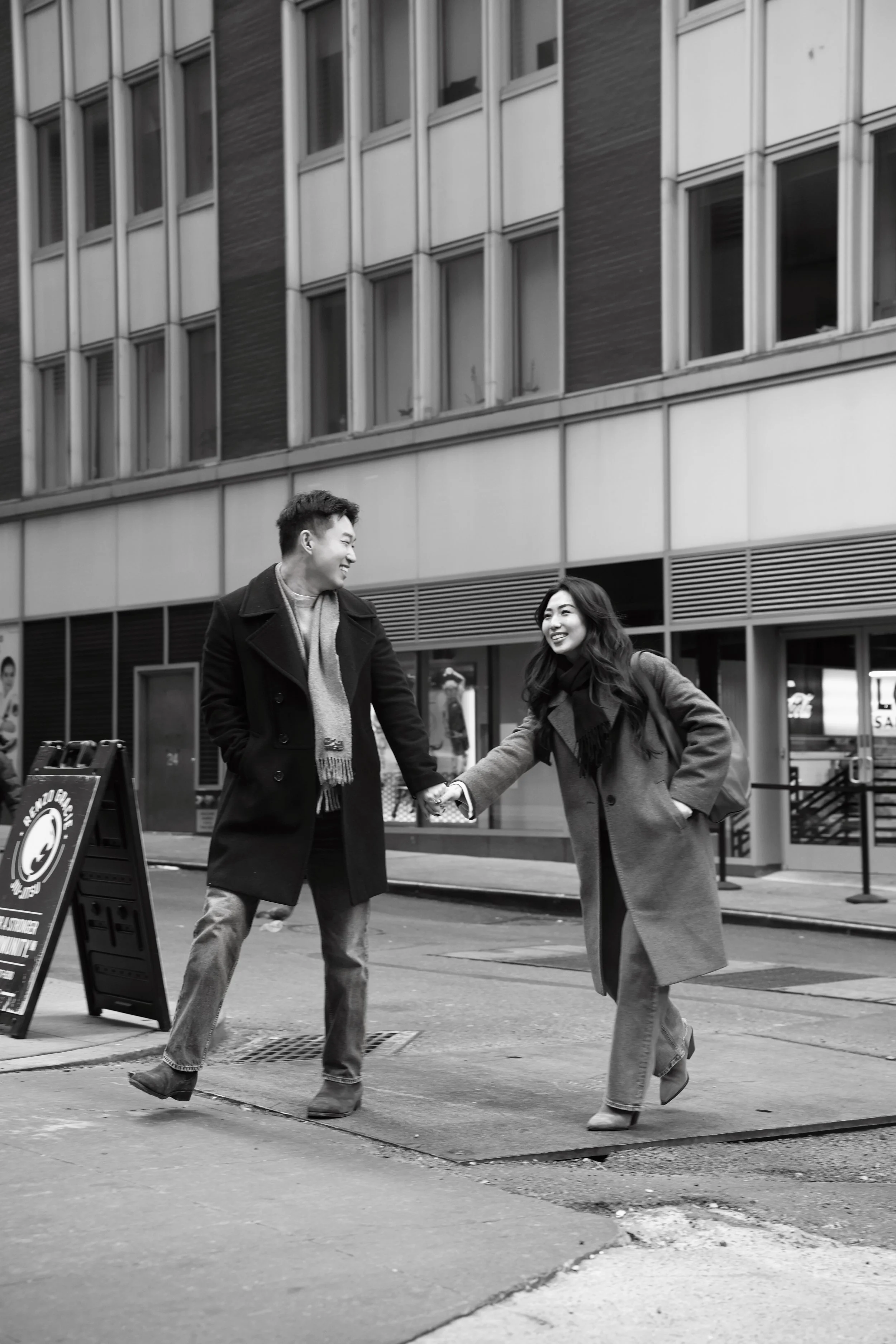 A black-and-white photo of a man and woman smiling and holding hands while crossing the street in an urban area, both wearing coats.