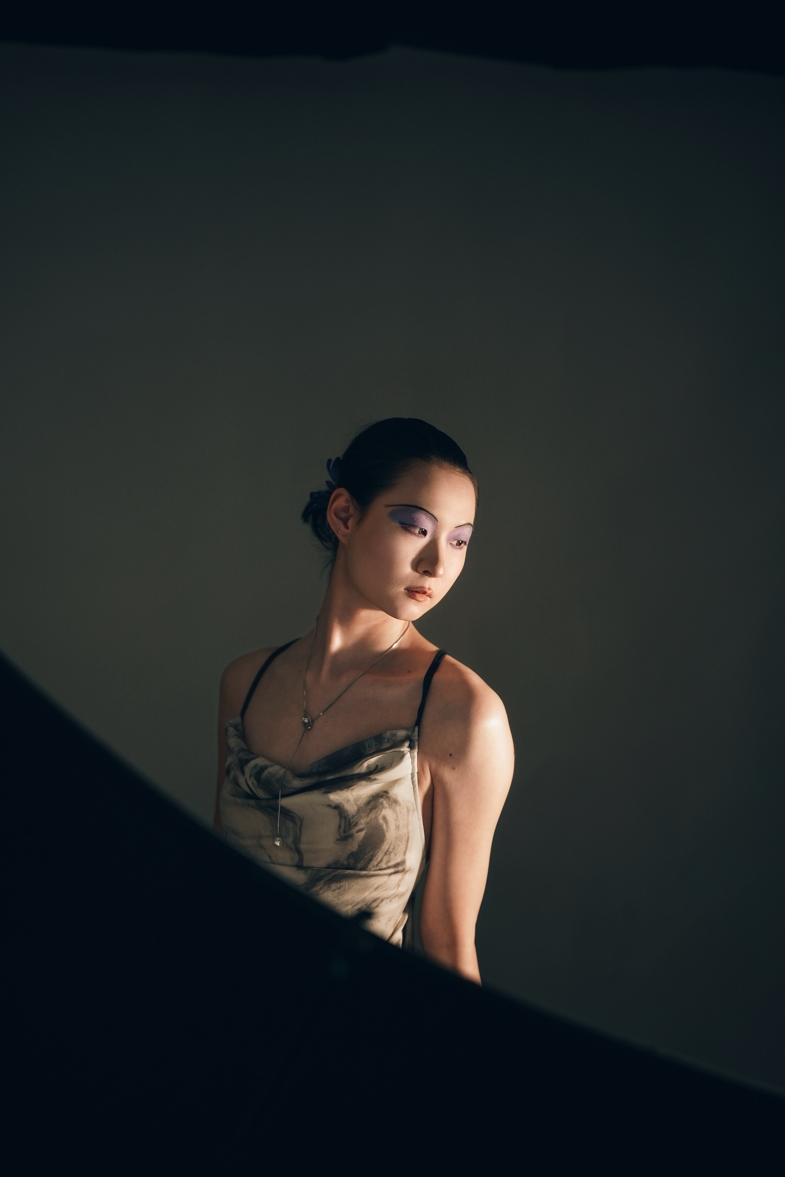 A woman with dark hair styled in an updo, wearing makeup with purple eyeshadow, a marble-patterned satin slip dress, and a necklace, standing against a plain, dark background and looking downward.