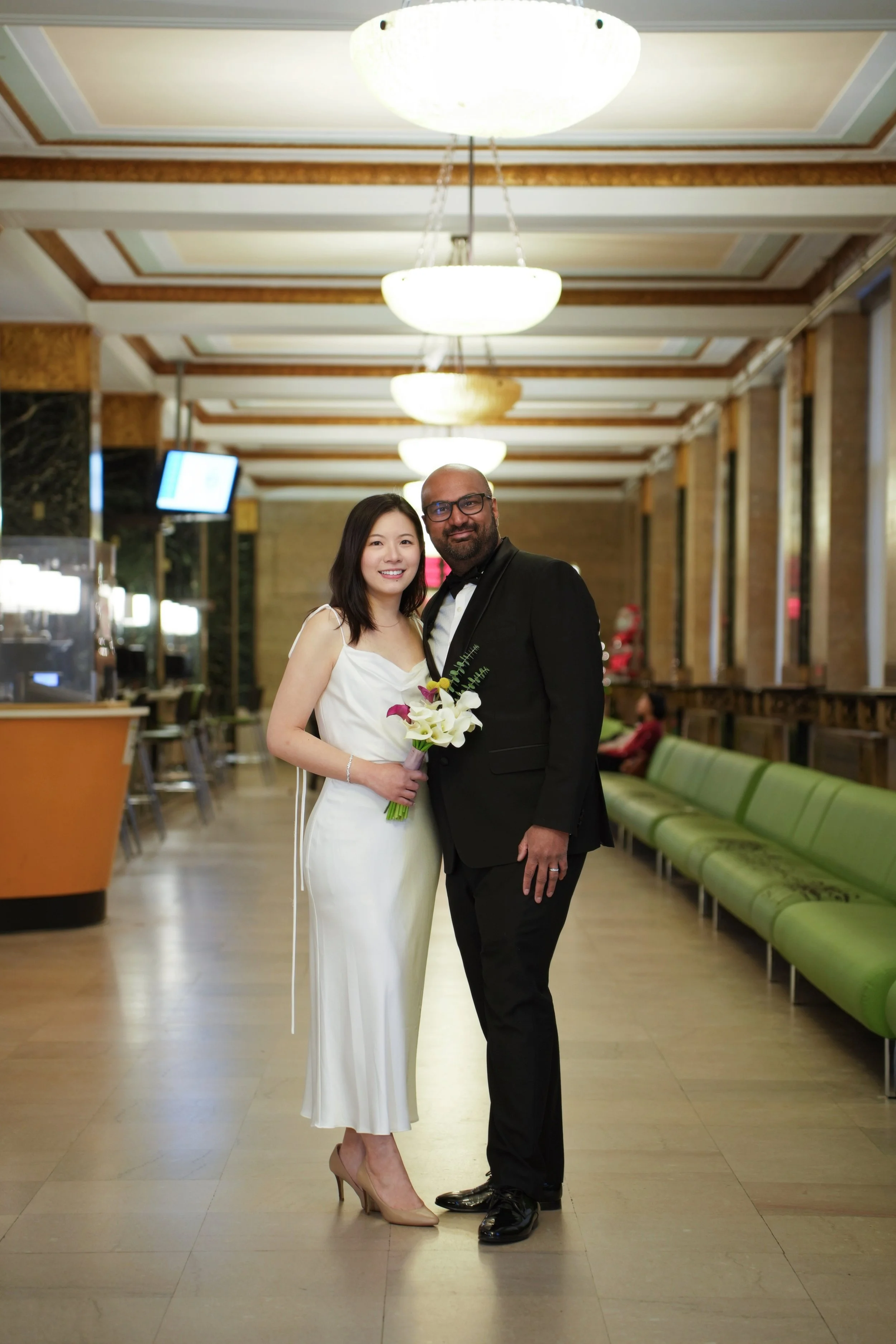 A man and a woman dressed in formal attire, standing together in an elegant indoor setting. The woman is holding a bouquet of flowers and wearing a white dress, and the man is in a black tuxedo with a bow tie. They are smiling and posing for the came