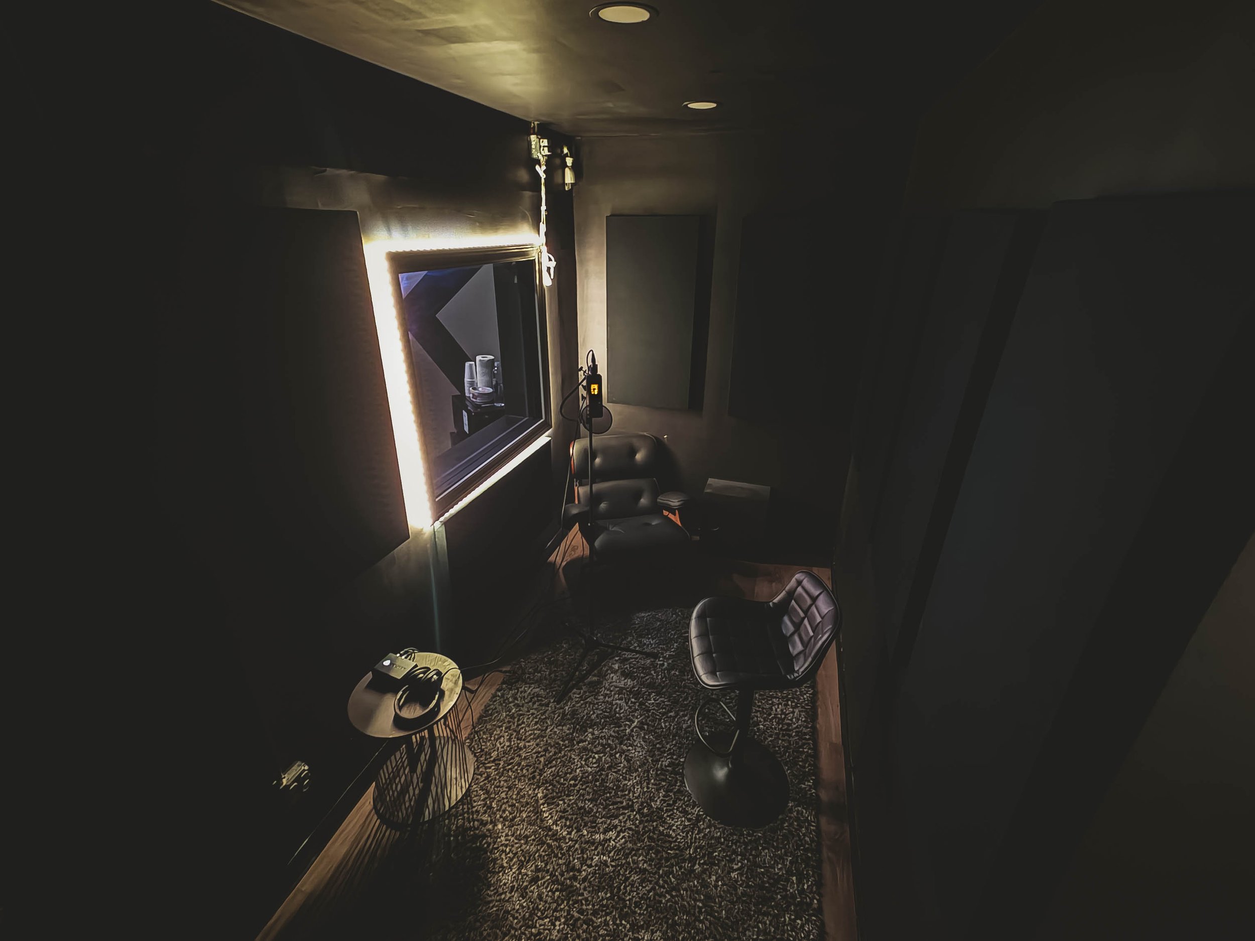 A dimly lit recording studio room with a leather chair, a small table with headphones, a patterned rug, and acoustic wall panels, with a window showing a music setup.