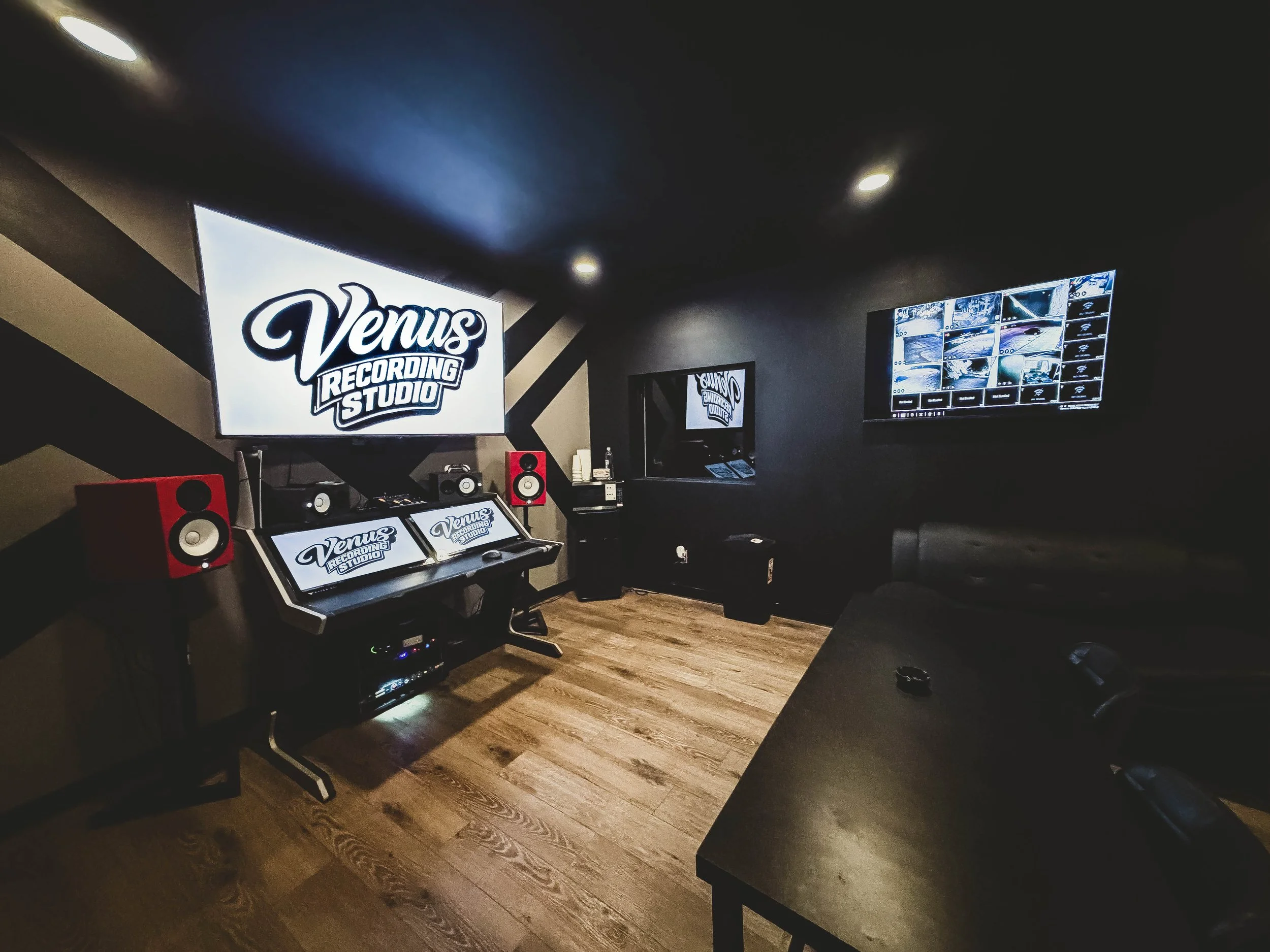 A recording studio room with a large screen displaying 'Venus Recording Studio' logo, multiple monitors, and red studio speakers, with dark walls and wooden floor.