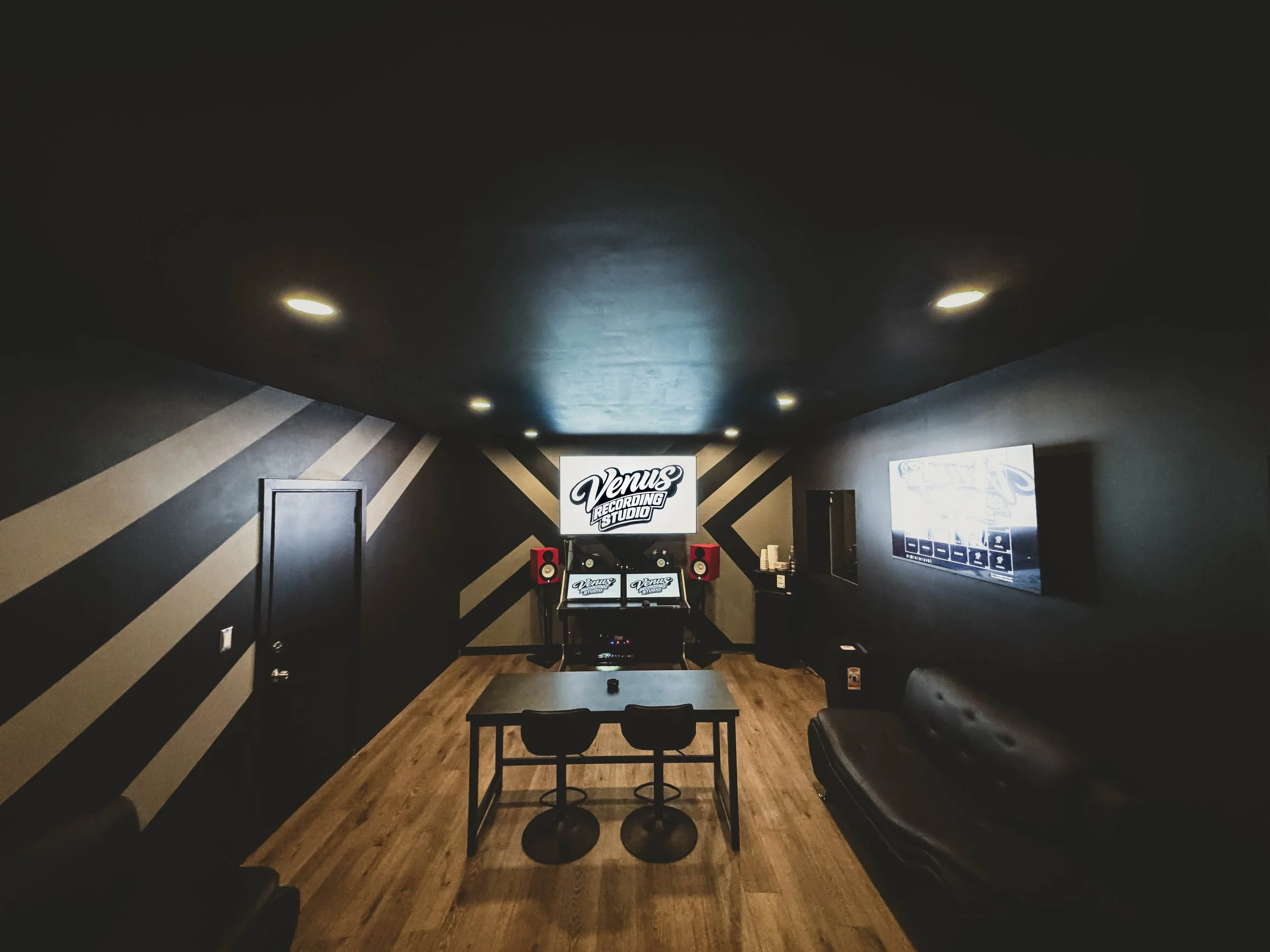 A modern recording studio with black walls, ceiling and couches. A large screen at the back displays 'Venus Recording Studio' and a TV on the right shows music software. There are two chairs at a table in the center, a pair of red speakers flanking the screen, and wood flooring.