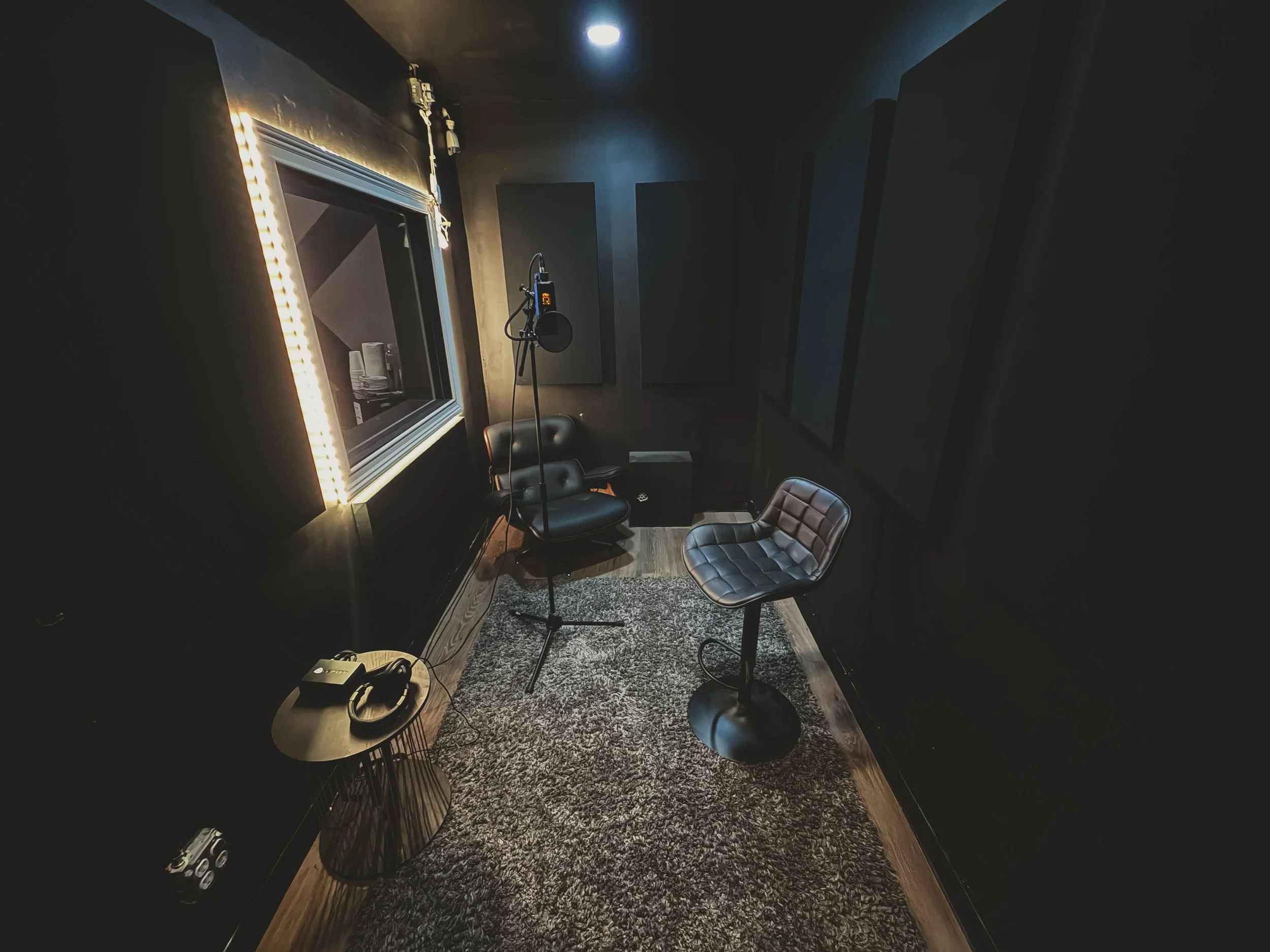 A recording studio with black walls, a large window with LED lights, a microphone on a stand, two black chairs, a small side table with headphones and a phone, and soundproofing panels on the walls.