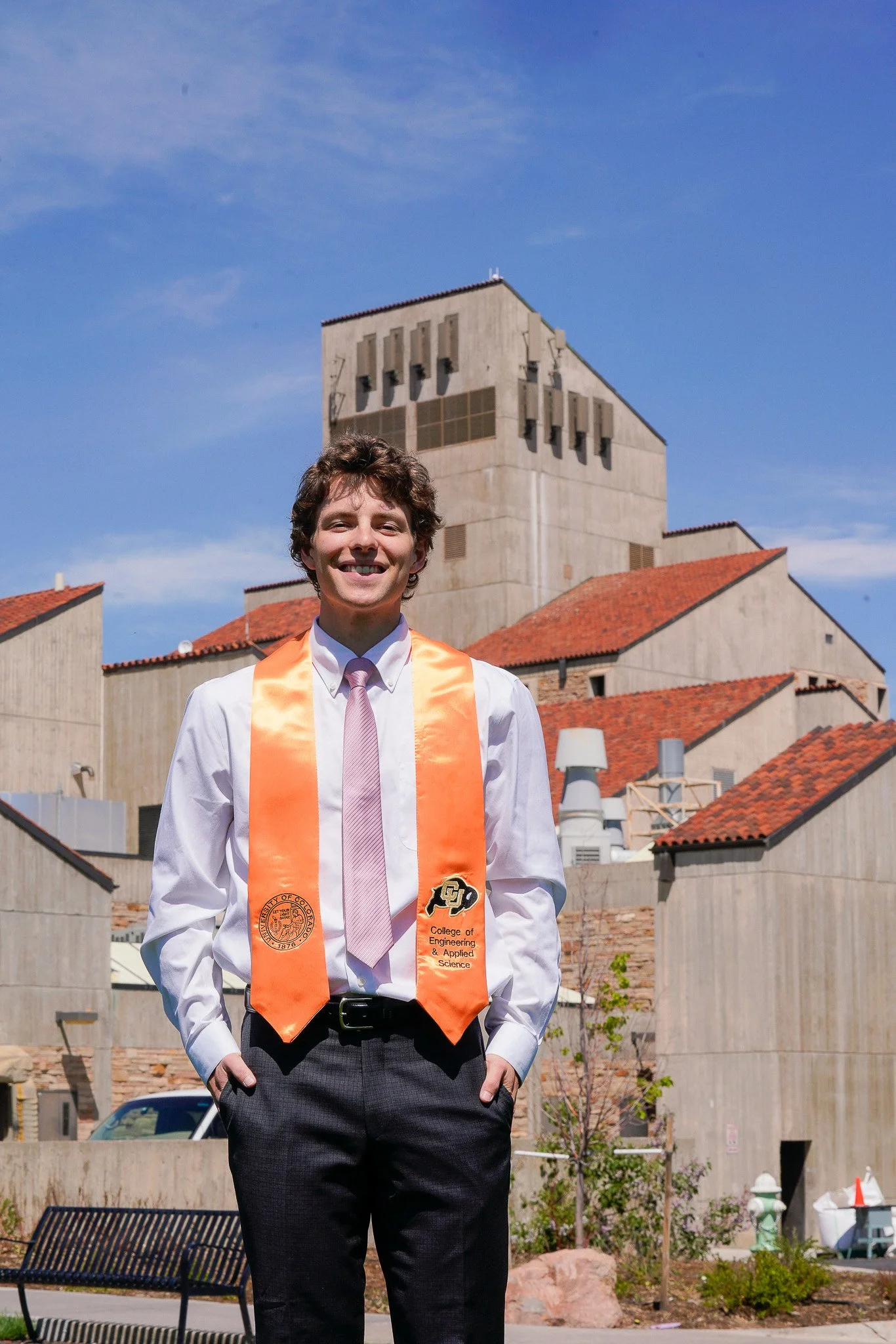 Young man in a white shirt, pink tie, and orange graduation stole standing outdoors with a building that has a concrete exterior and red tile roof behind him. Graduation photo taken at the University of Colorado Boulder (CU Boulder).
