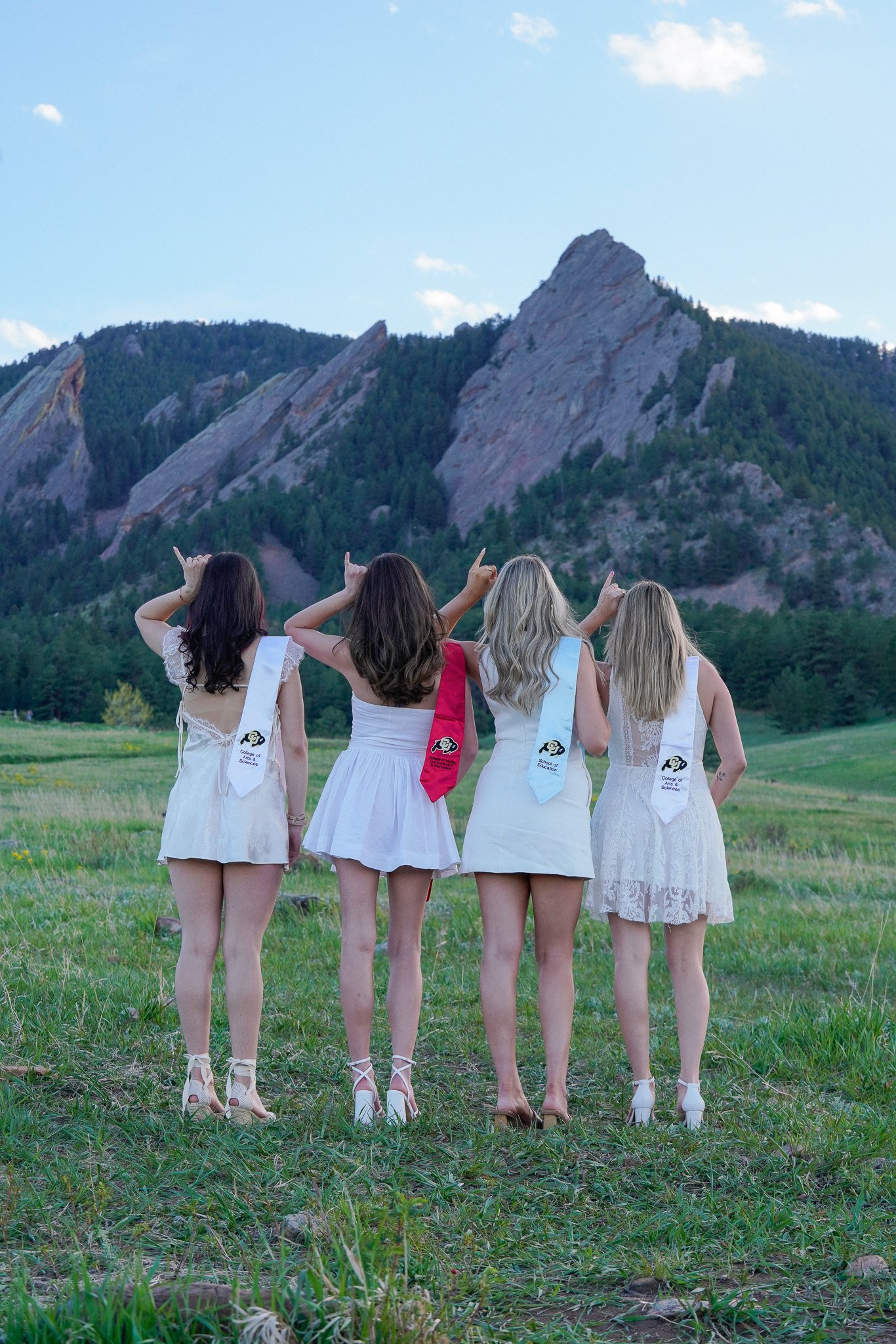 Four women dressed in white dresses and graduation stoles, standing in a grassy field, pointing towards a mountain range in the distance. Graduation photo located in front of the Flatiorns at Chautauqua Park.