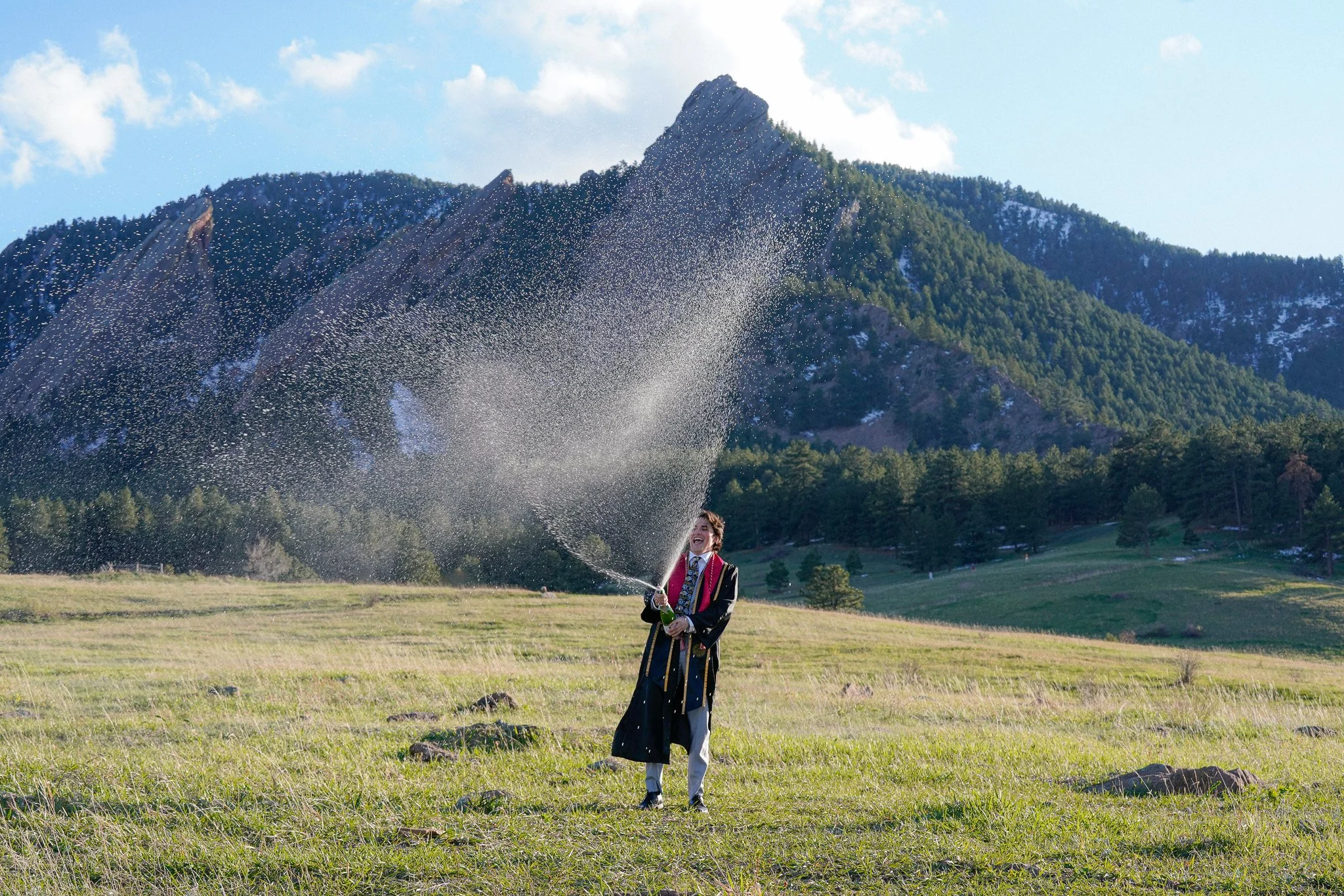 A person in academic regalia celebrating by spraying champagne in an open field with mountains and trees in the background. Graduation photo located in front of the Flatiorns at Chautauqua Park.
