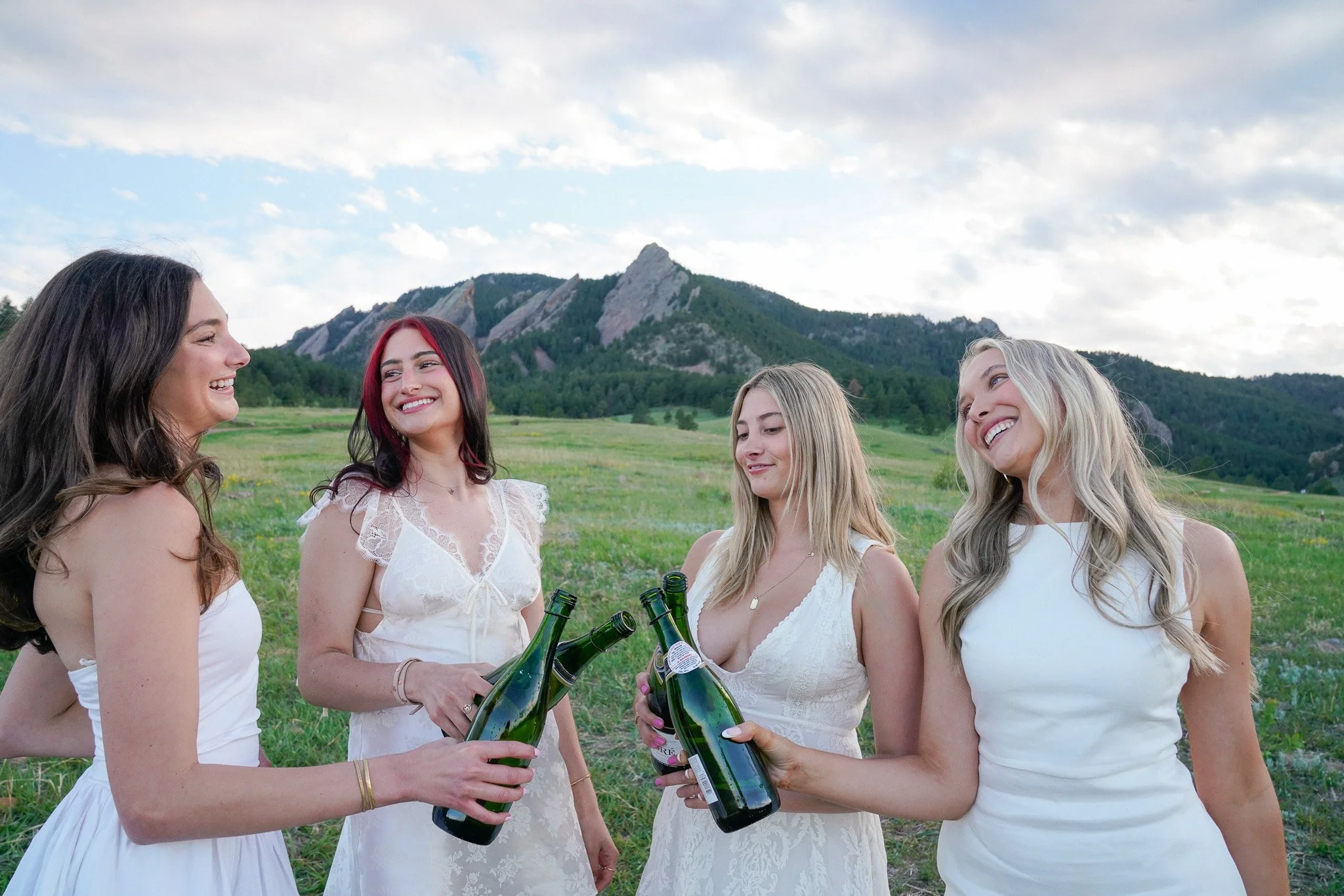 Four women in white dresses holding beer bottles, smiling and talking outdoors in a green field with mountains in the background. Graduation photo located in front of the Flatiorns at Chautauqua Park.